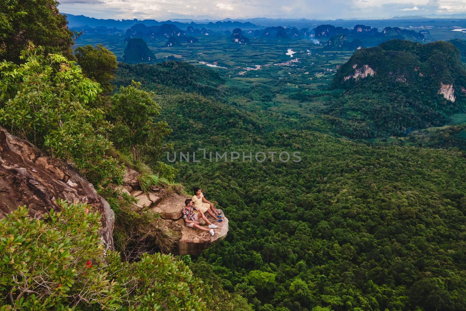 Dragon Crest Mountain Krabi Thailand, a Young couple sits on a rock that overhangs the abyss with a beautiful landscape. Dragon Crest or Khuan Sai at Khao Ngon Nak Nature Trail in Krabi rainforest