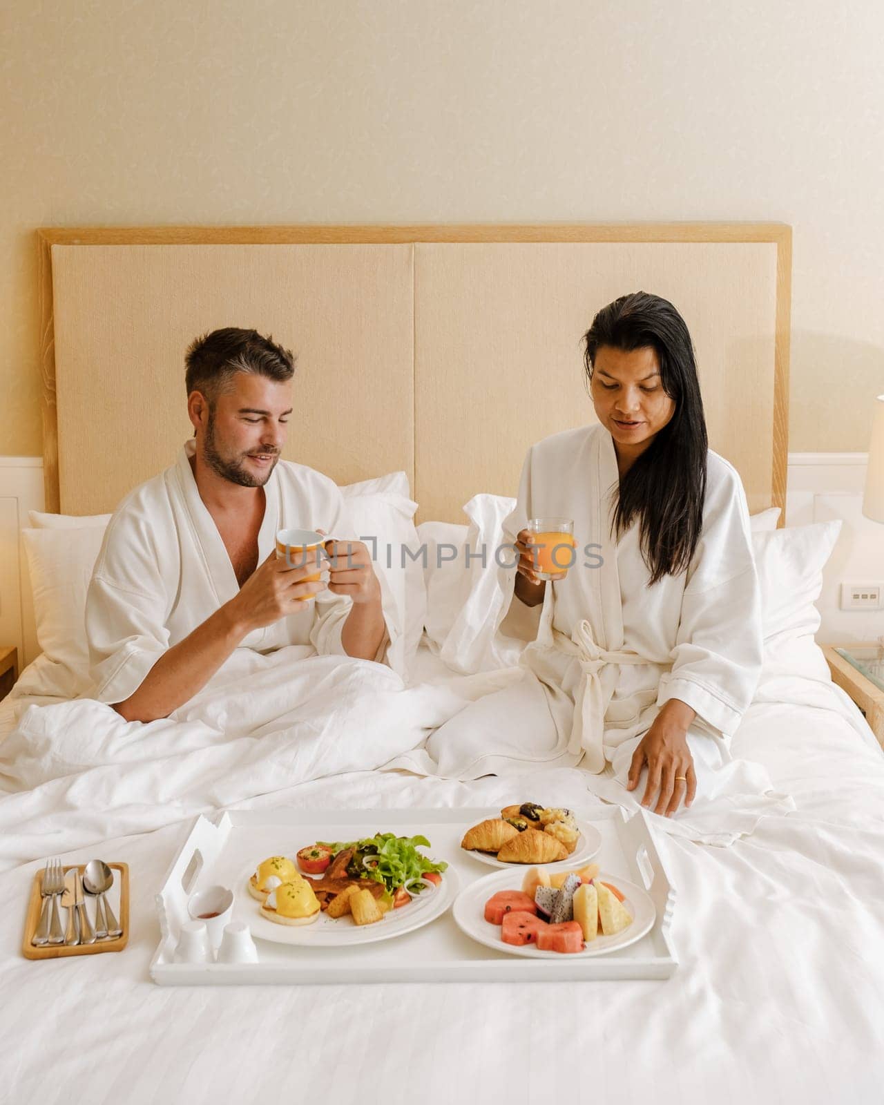 couple having breakfast in bed at a luxury hotel, men and women in bed with breakfast coffee, and bread. a diverse couple of an Asian woman and a European man in bedroom