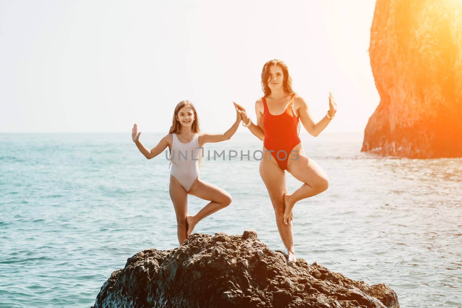 Woman and her daughter practicing balancing yoga pose on one leg up together on rock in the sea. Silhouette mother and daughter doing yoga at beach by panophotograph