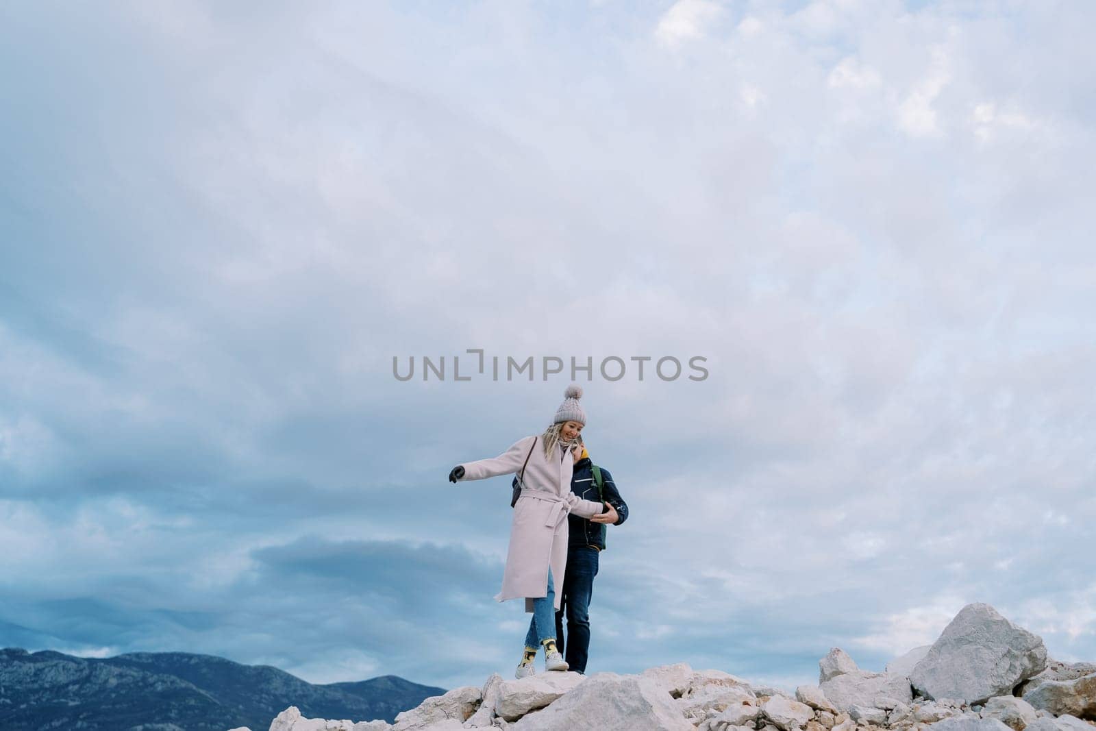 Man helps woman walk on the rocks, holding her hand in the mountains by Nadtochiy