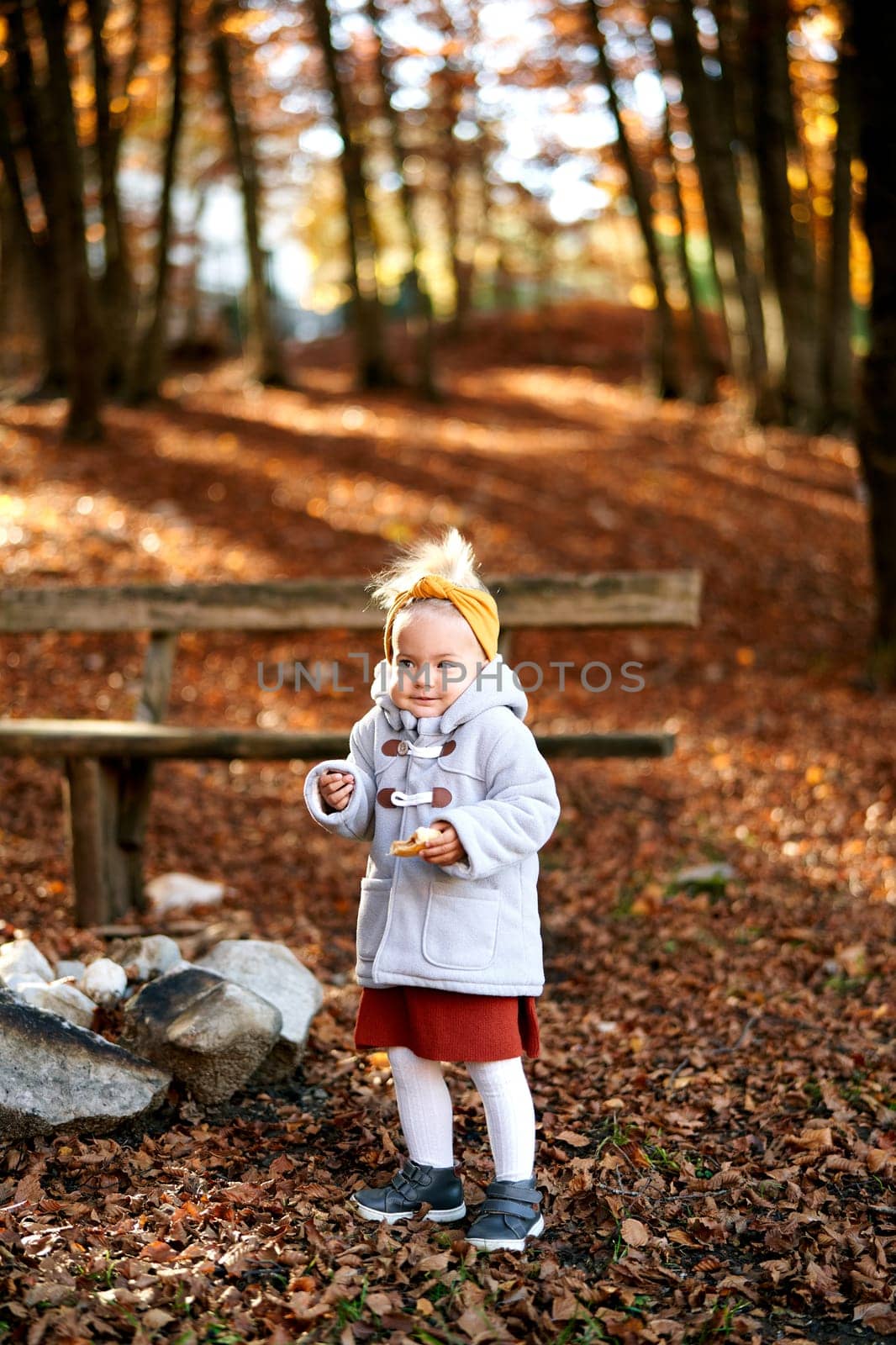 Little girl with a bun in her hand shrugs her shoulders while standing near a bench in the park by Nadtochiy