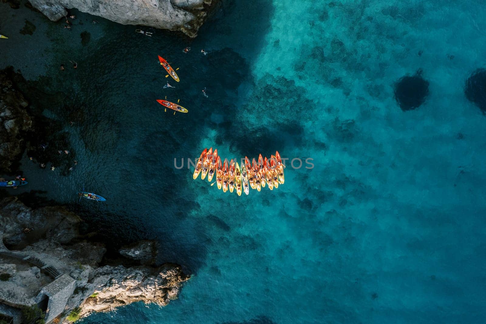 Row of red and yellow kayaks sits in a cove near the rocky shore. Drone by Nadtochiy