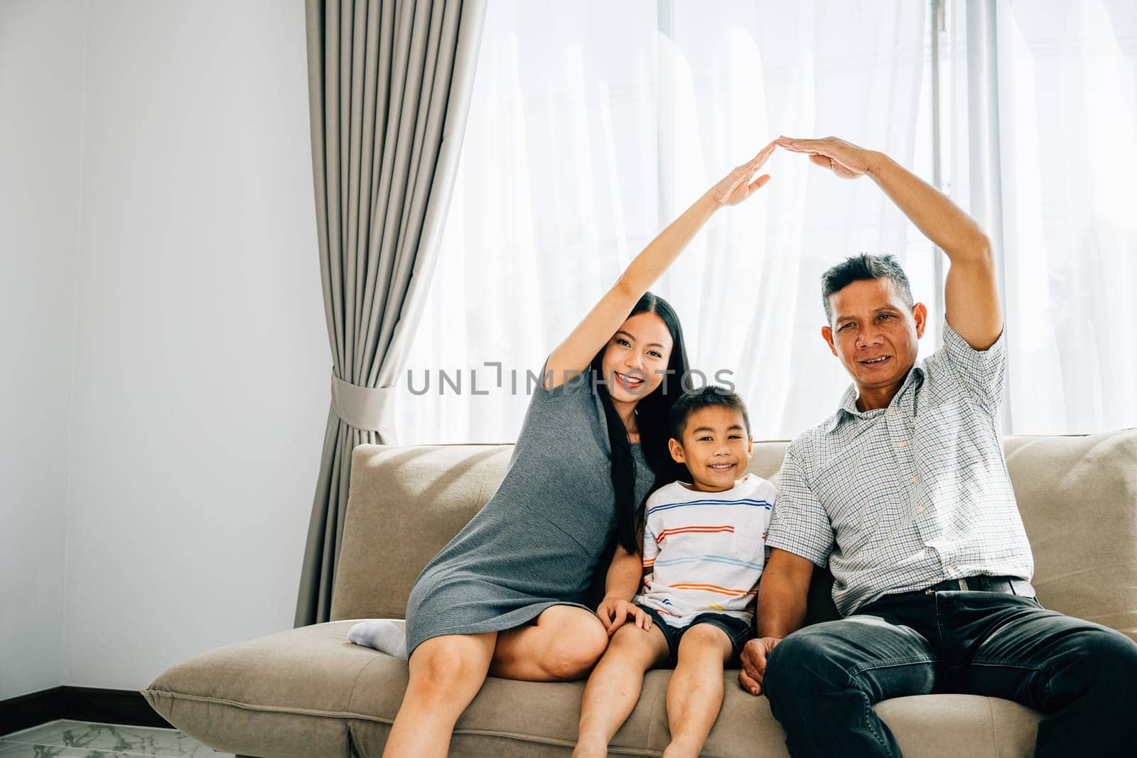 A joyful family poses on a sofa parents making a roof gesture above their little son by Sorapop