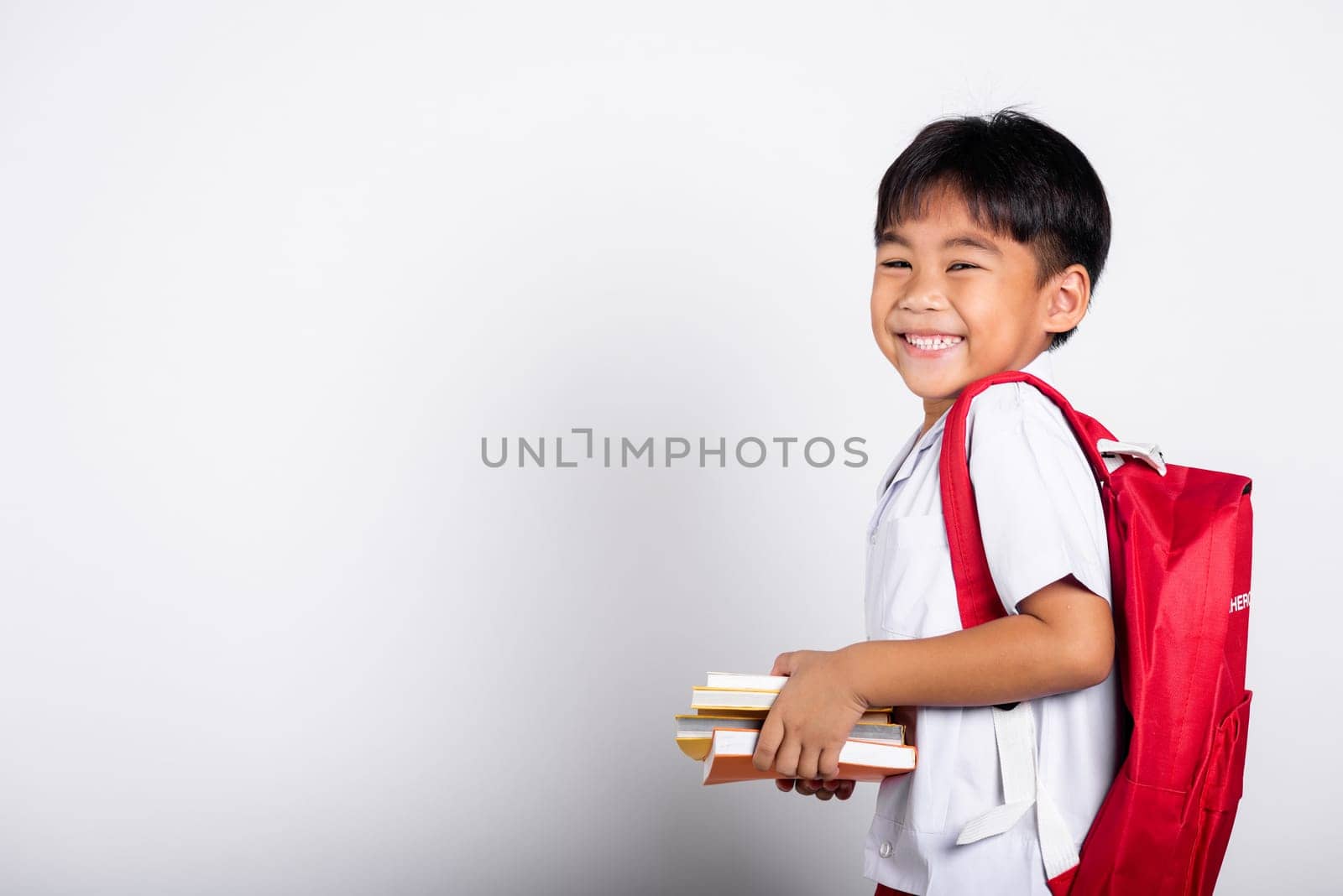 Asian adorable toddler smiling happy wearing student thai uniform red pants stand books for study ready for school by Sorapop