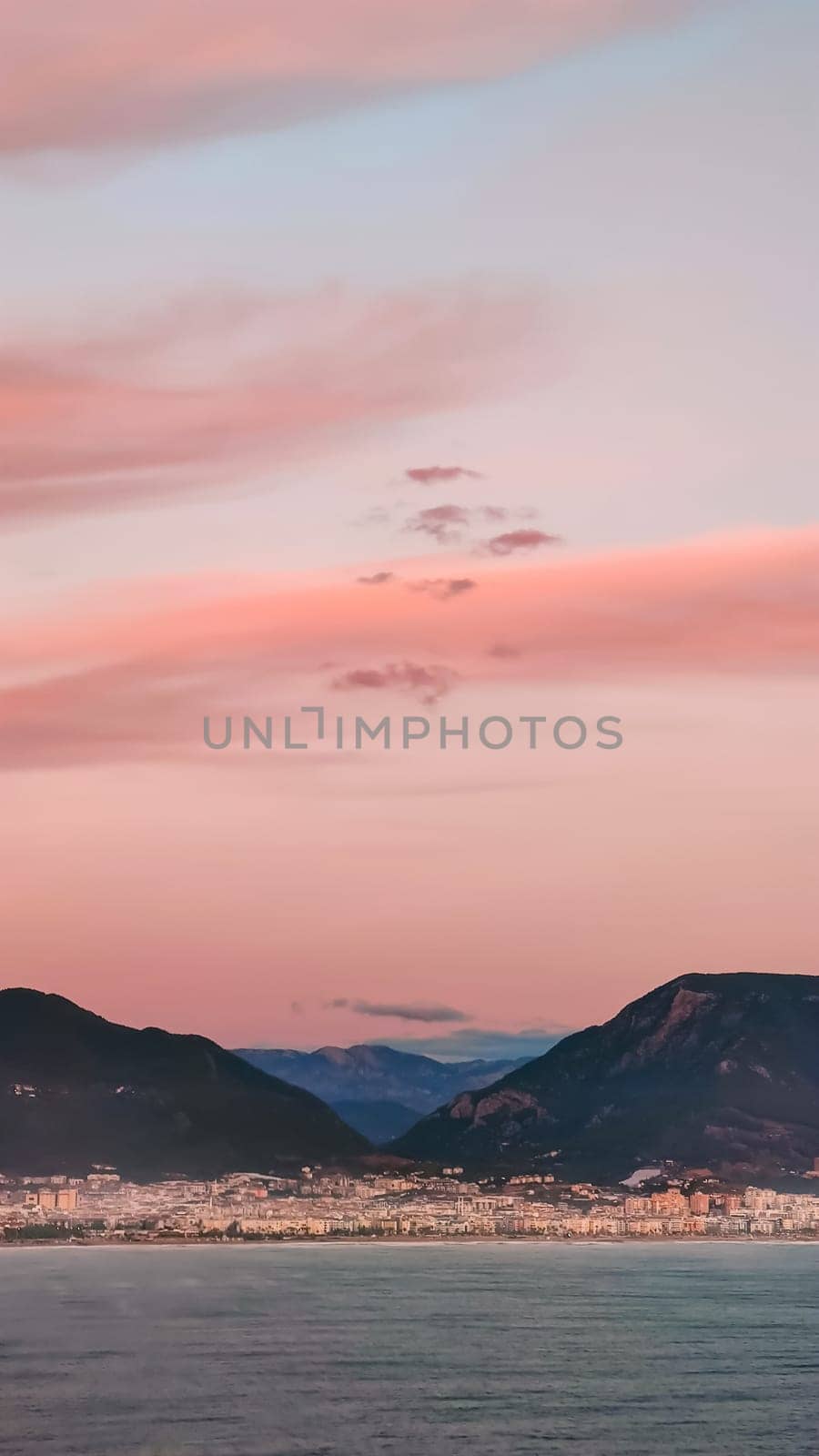 Pink clouds in evening sky over blue sea on background of city and mountains on horizon, vertical frame.