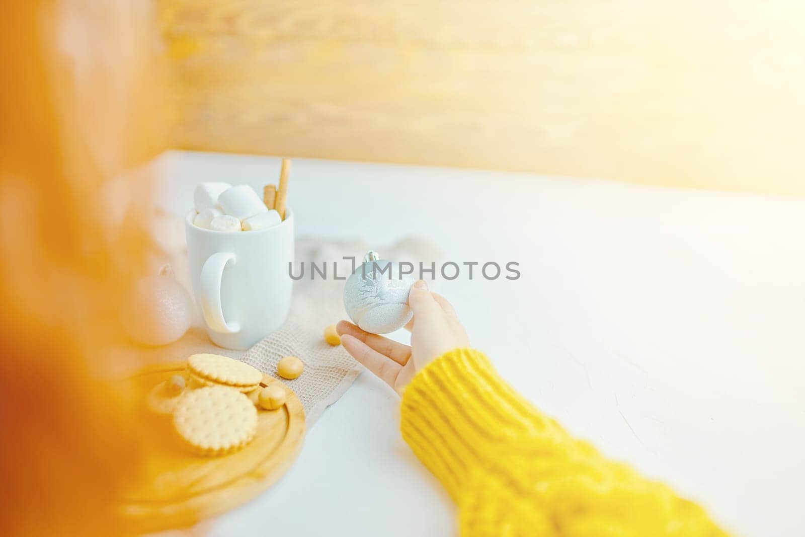 Woman in a yellow sweater drinks hot cocoa with marshmallow and cinnamon rolls in a white mug and cookies and a figure of a Christmas tree