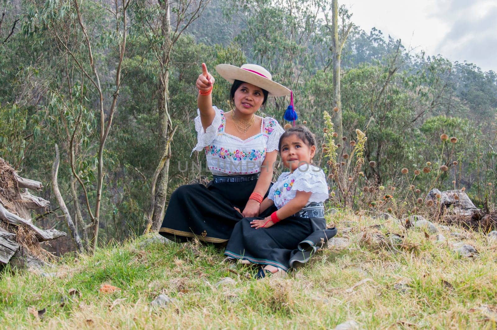 mother pointing her hand upward while her daughter looks on curiously. they are dressed in traditional dresses from otavalo, ecuador in the jungle. women's day. by Raulmartin
