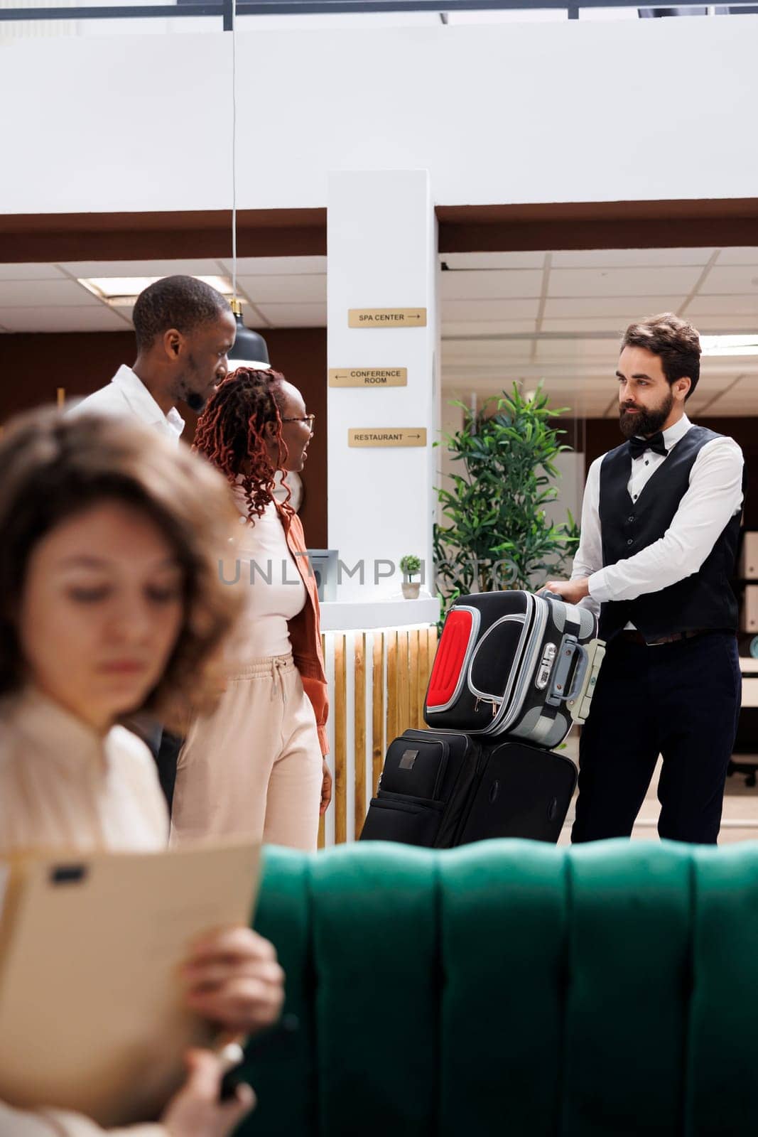 Male employee helping clients with bags, carrying luggage for people after doing check in procedure. Young couple receiving assistance from luxury resort bellboy at reception desk.