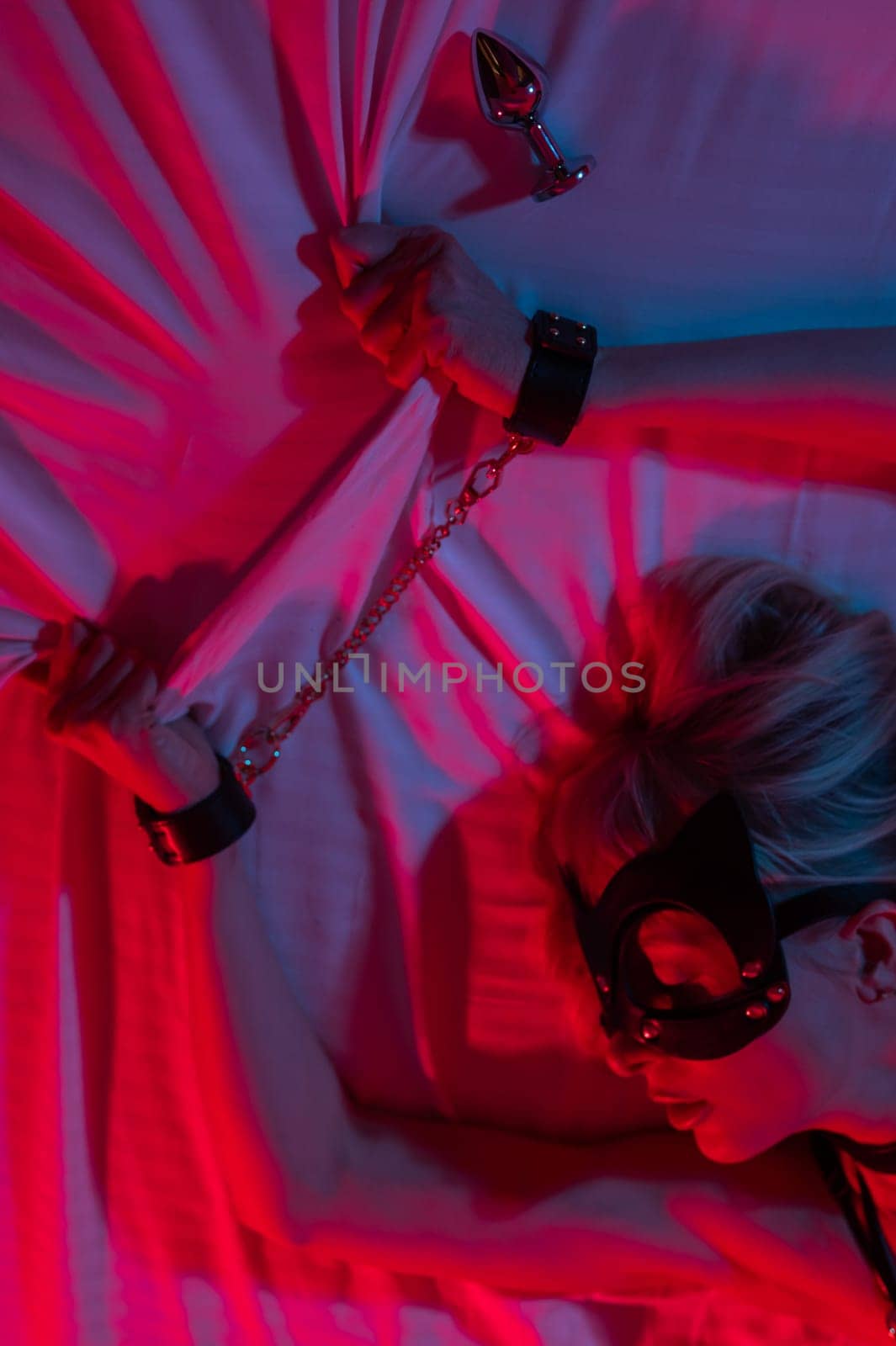 Sexy blonde woman in a leather mask and handcuffs and a collar lies in the bedroom in a red-blue neon light. Role-playing games