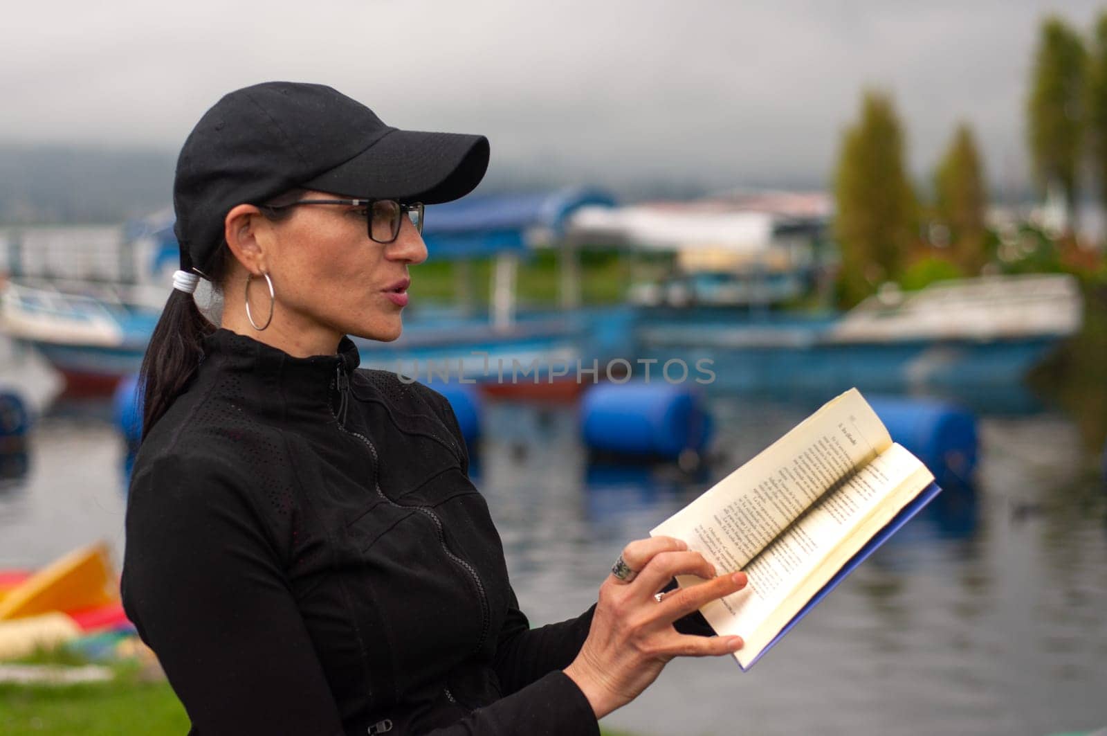 woman in her forties in black clothes reading a book on a pier with boats in the background. book day. High quality photo