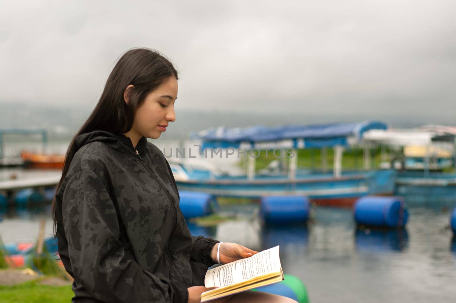 attractive young man from latin america sitting by a pier with boats reading a book while it rains. book day by Raulmartin