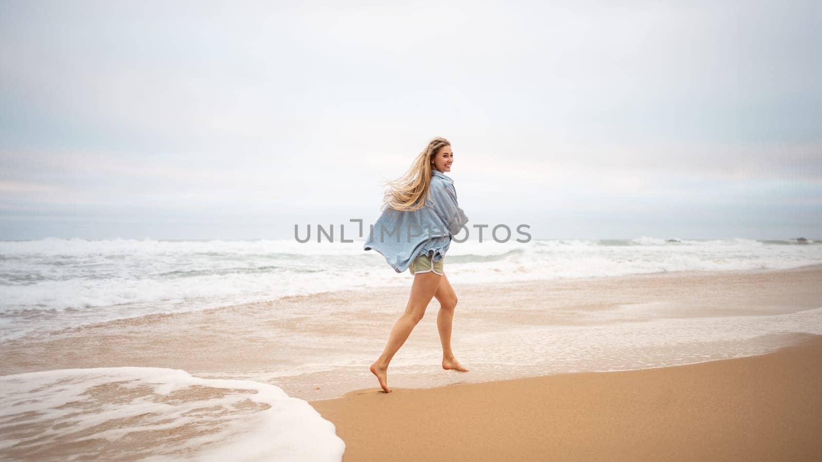 Woman running ocean beach and turns around, smiling at camera. Happy female traveler dressed denim shirt walk sandy sea beach, holiday vacation at summer.