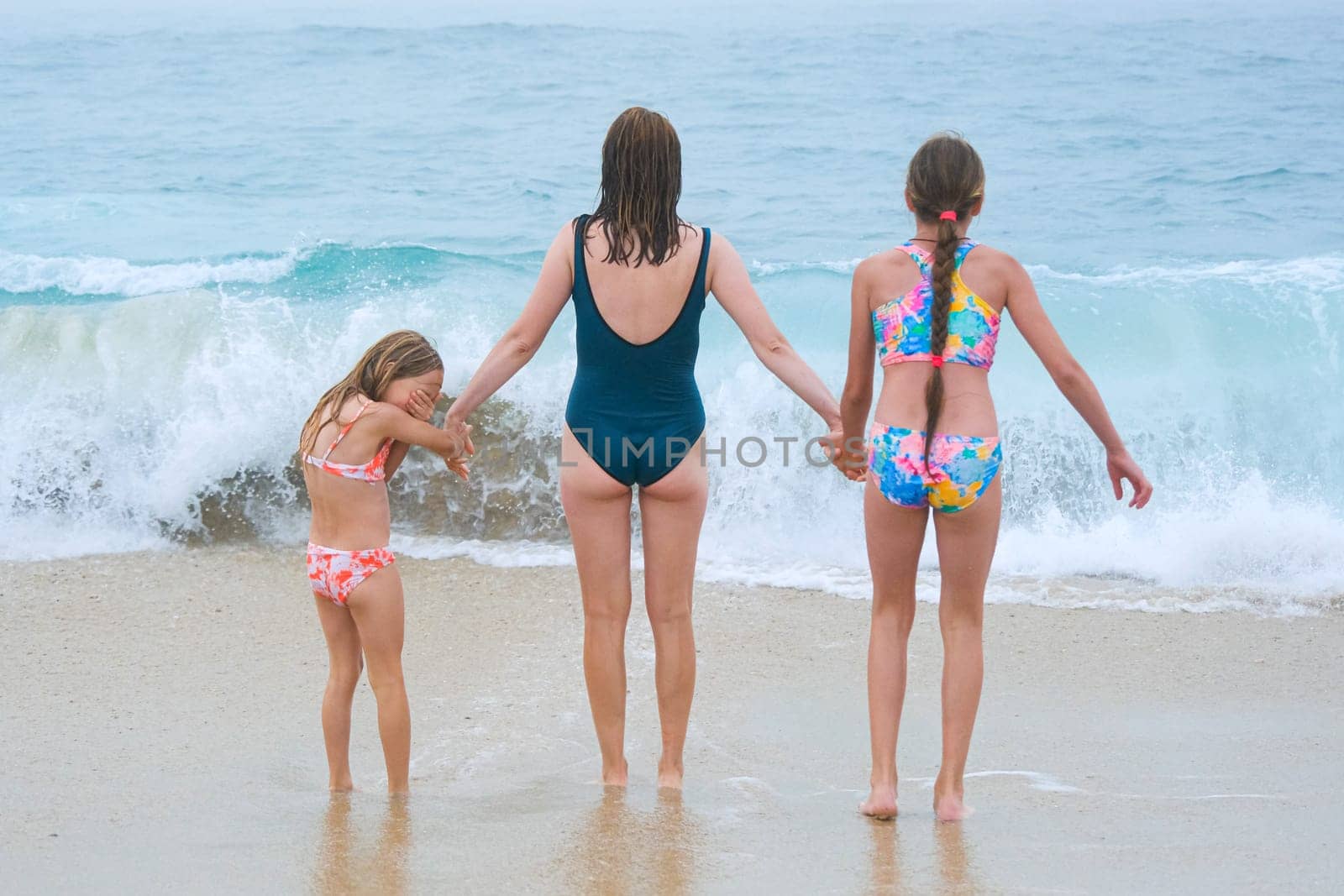 Mother and children playing on the ocean beach. Family enjoying the ocean. Mother holds girls's hands and they all look at the ocean together.