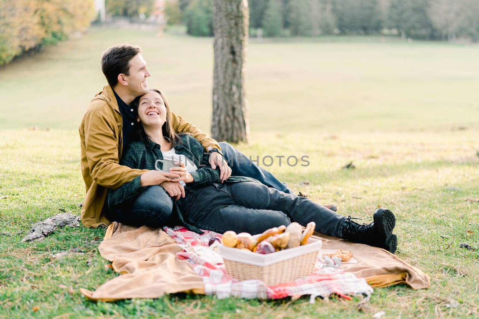 Smiling woman lying on blanket in park leaning against seated man by Nadtochiy
