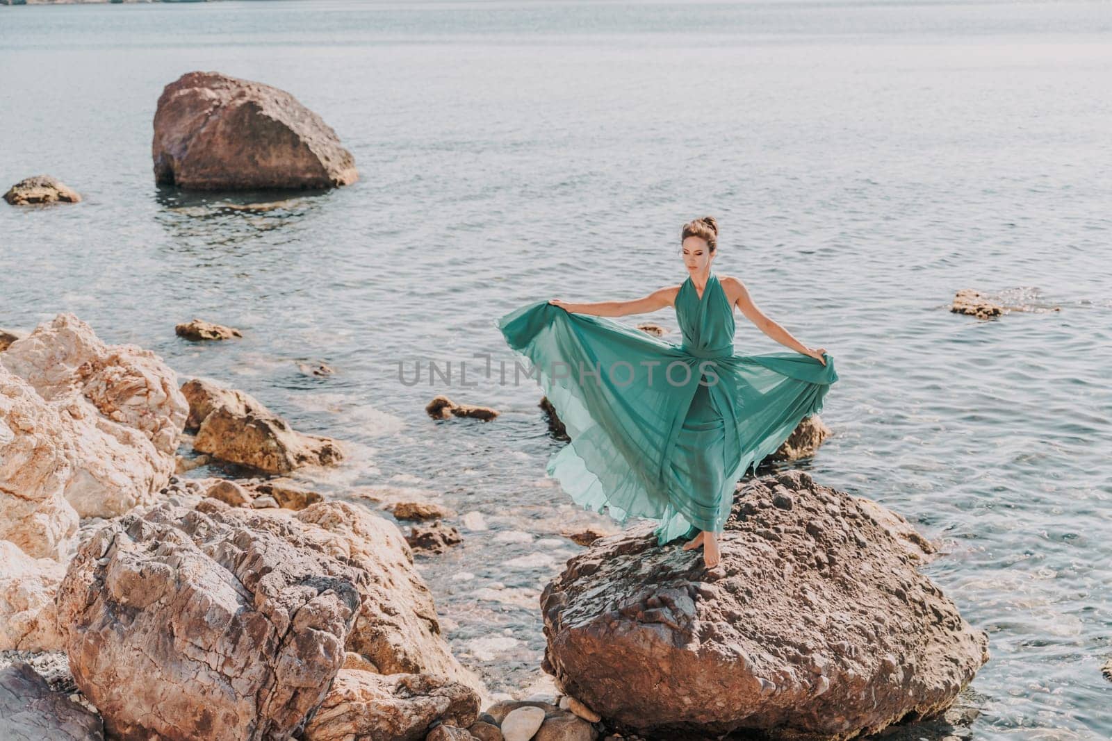 Woman green dress sea. Female dancer in a long mint dress posing on a beach with rocks on sunny day. Girl on the nature on blue sky background