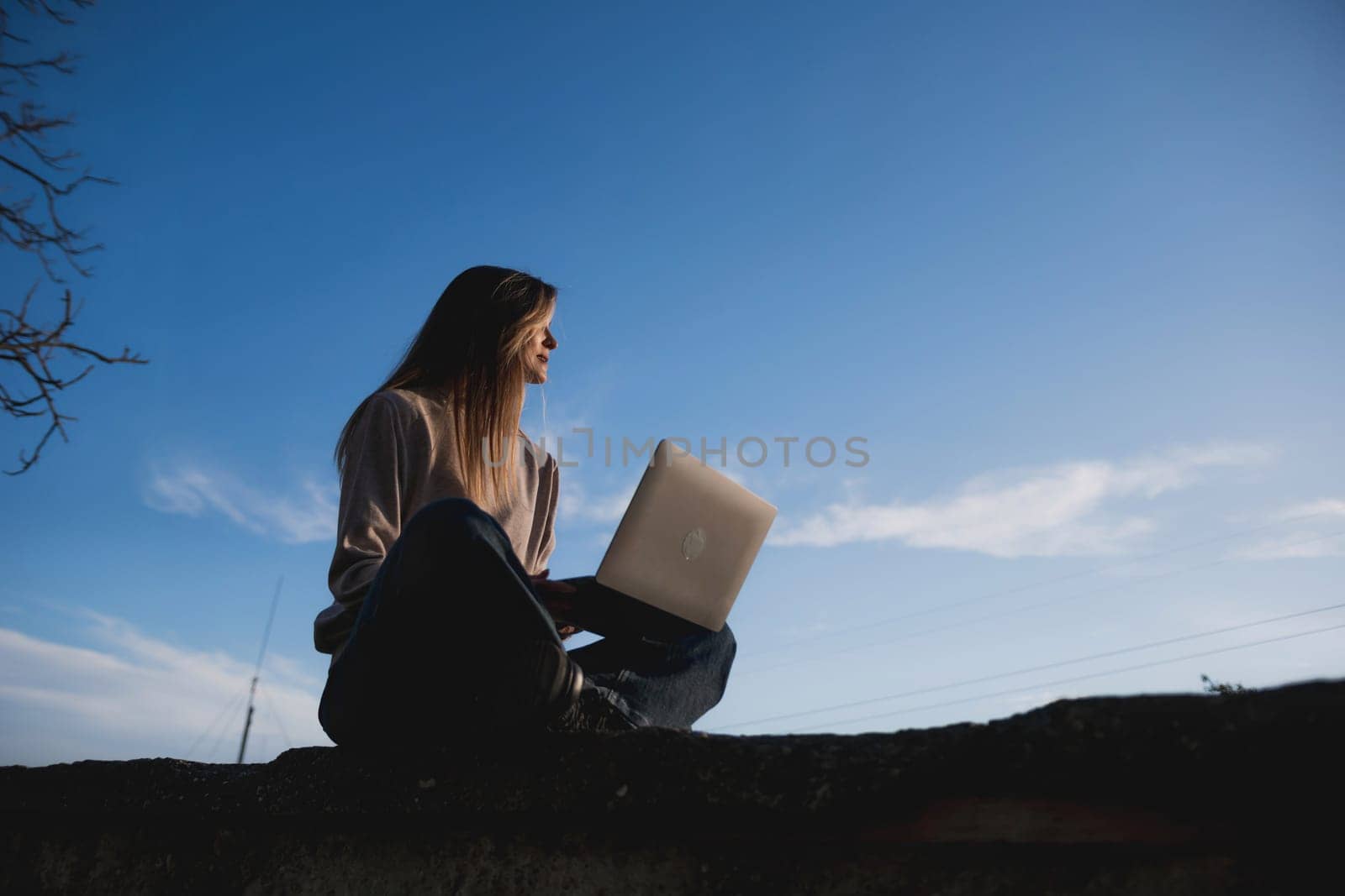 Woman freelancer uses laptop on cement wall outdoors against the sky. The woman to be focused on her work or enjoying some leisure time while using her laptop. by Matiunina