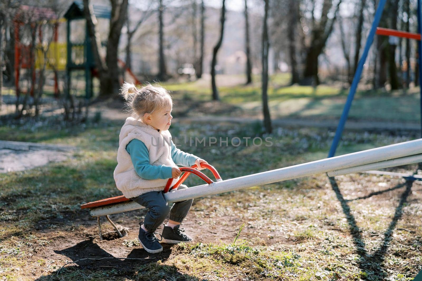 Little girl sits on a swing-balancer holding on to the handles in the playground. High quality photo