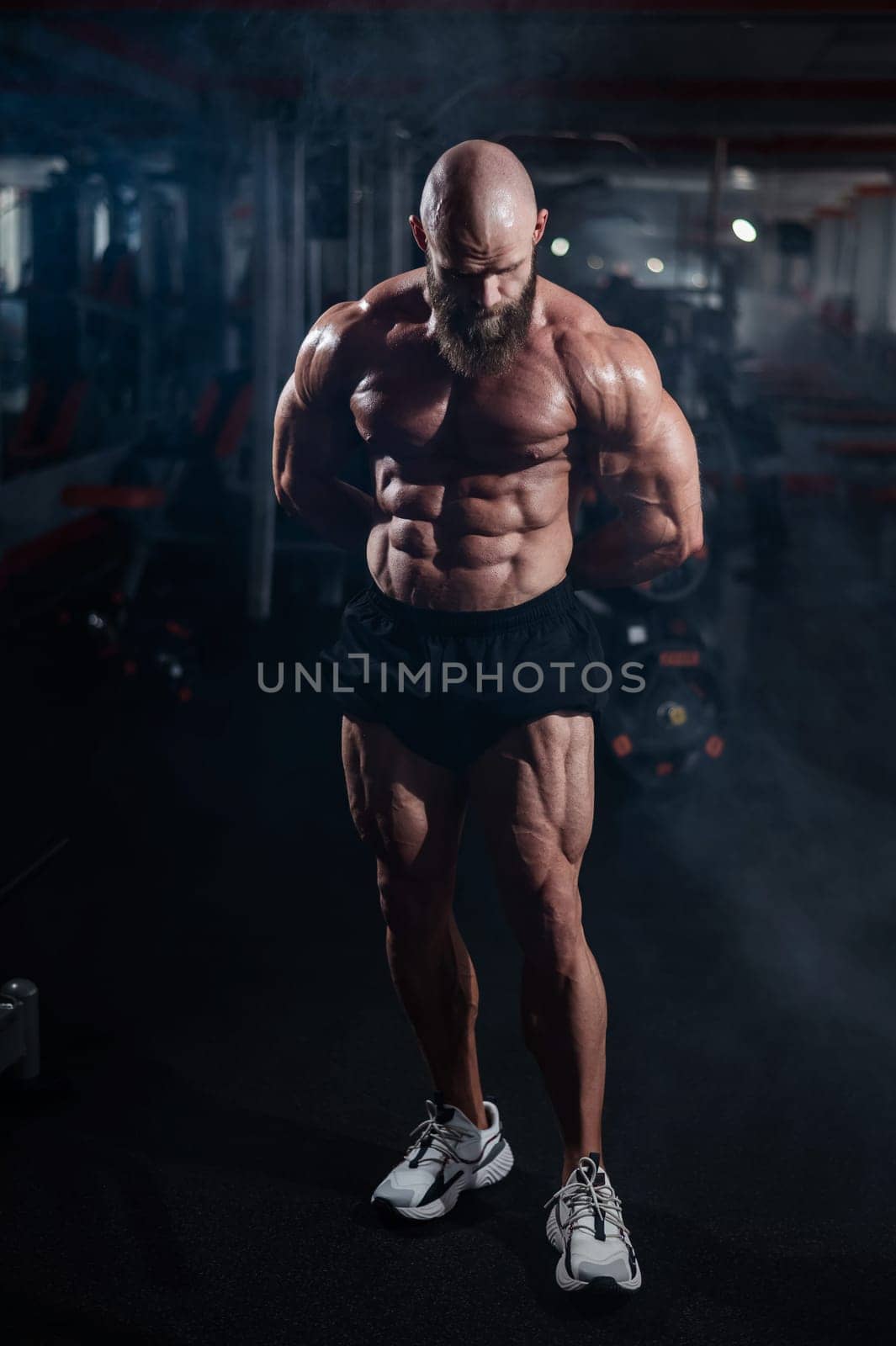 Muscular bald man posing in shorts. Bodybuilder showing off his shape in the gym