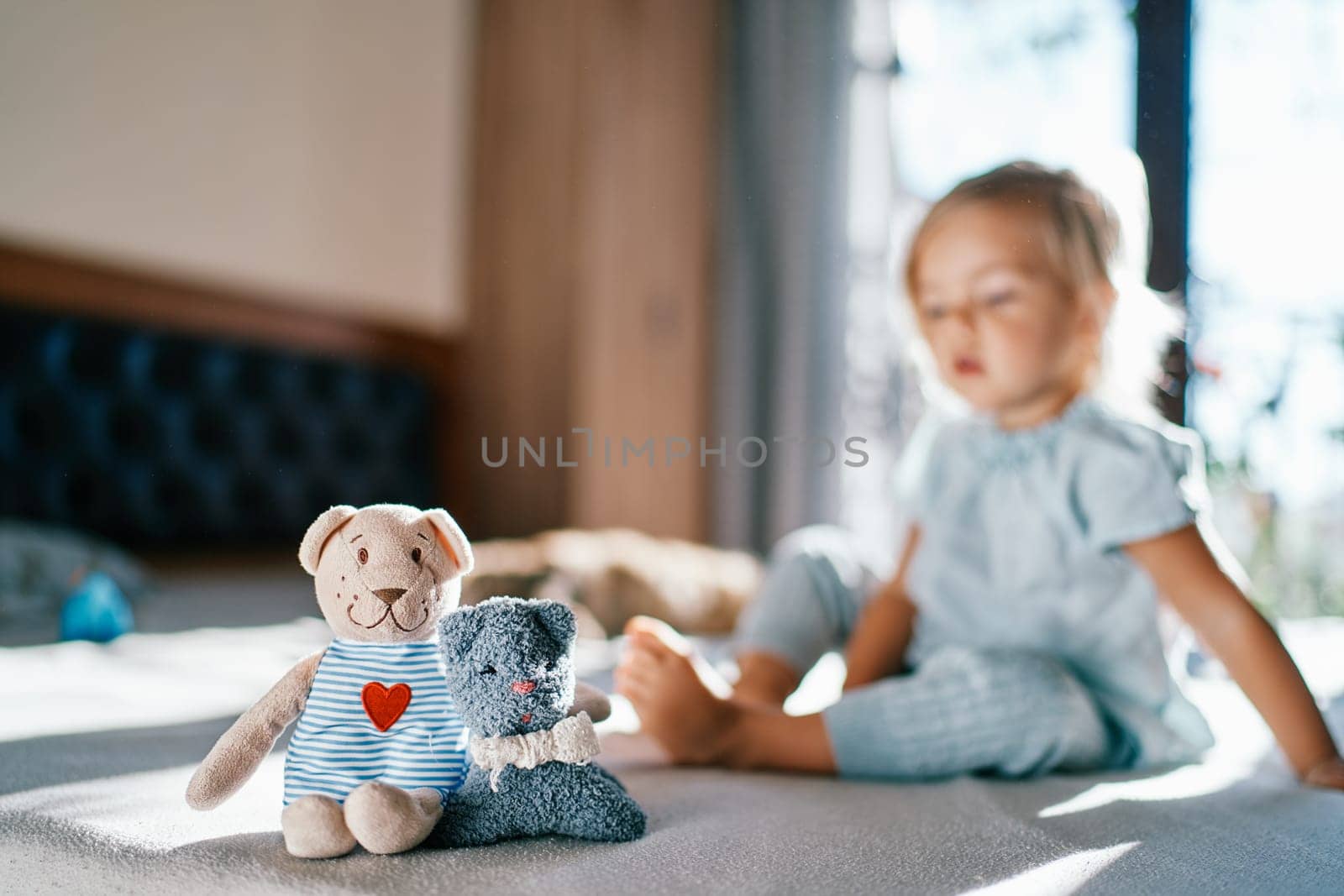 Teddy bear and a toy cat are sitting on the bed embracing against the background of a little girl by Nadtochiy