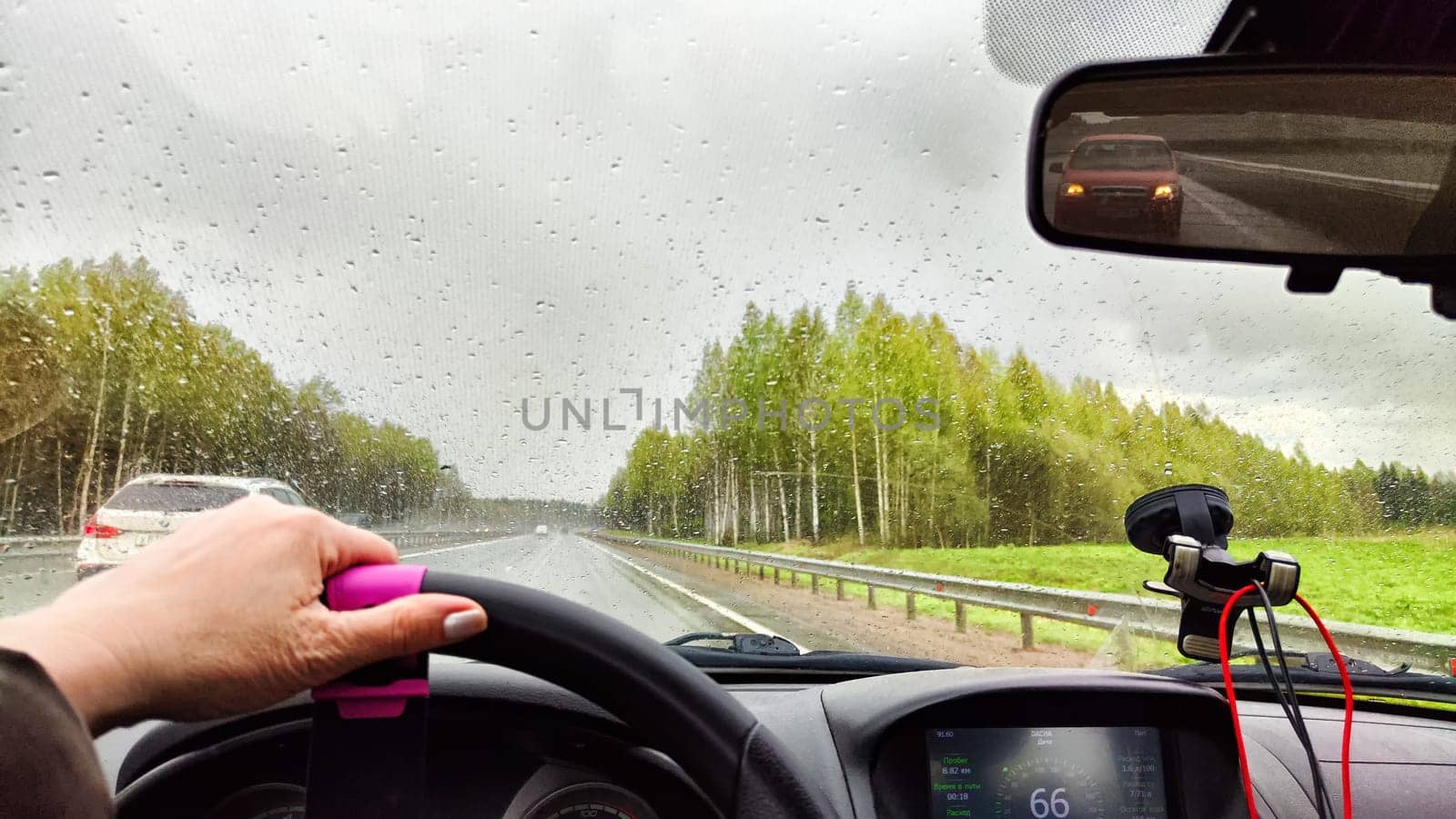 View from car windshield from the driver's seat and hand on the steering wheel of woman on during rain, drops on the glass and nature. Travel in rainy autumn, spring or summer weather