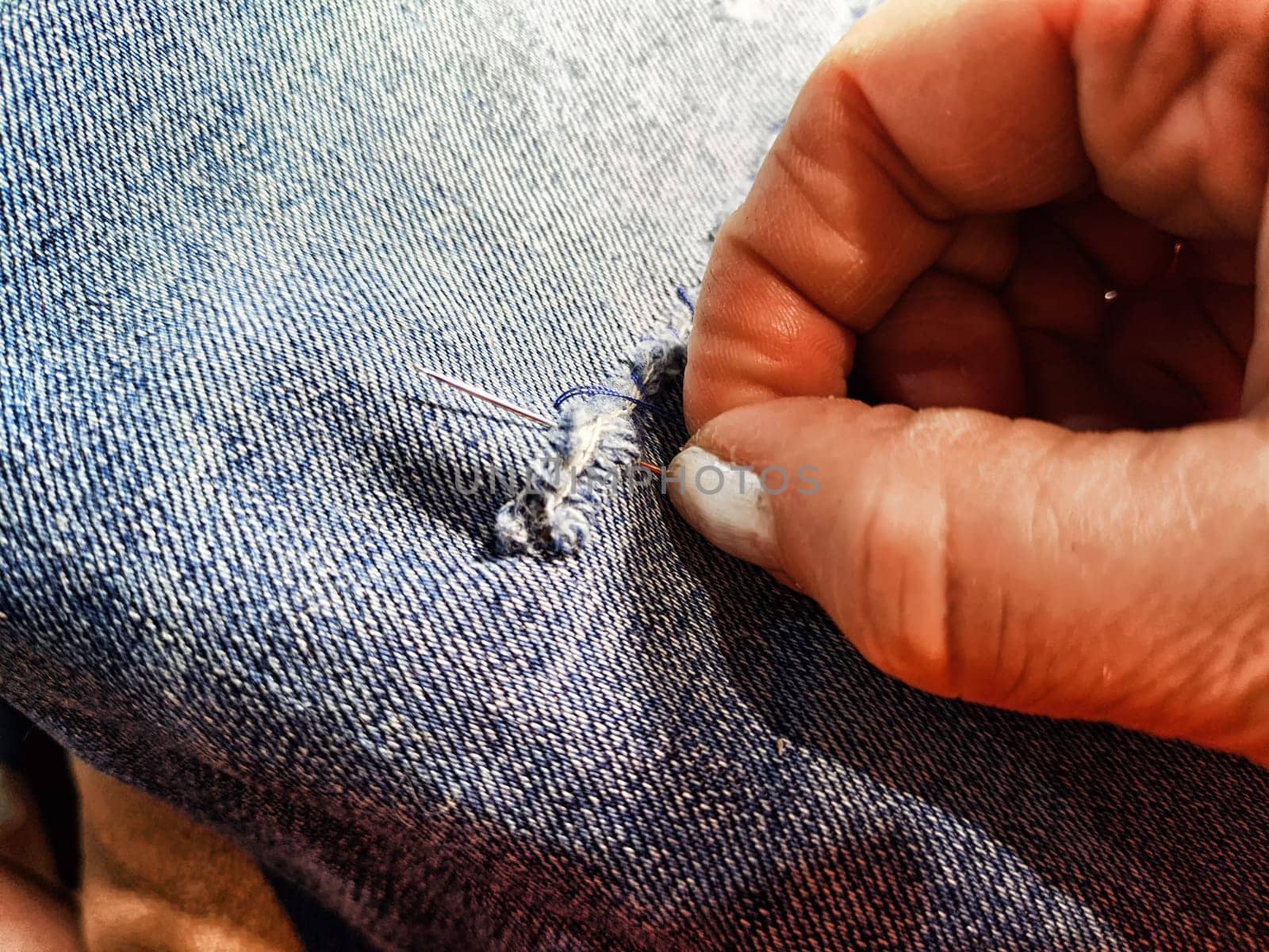 Woman repairing a pair of blue jeans. Fingers with a needle and torn jeans fabric. Sewing up a tear on blue fabric