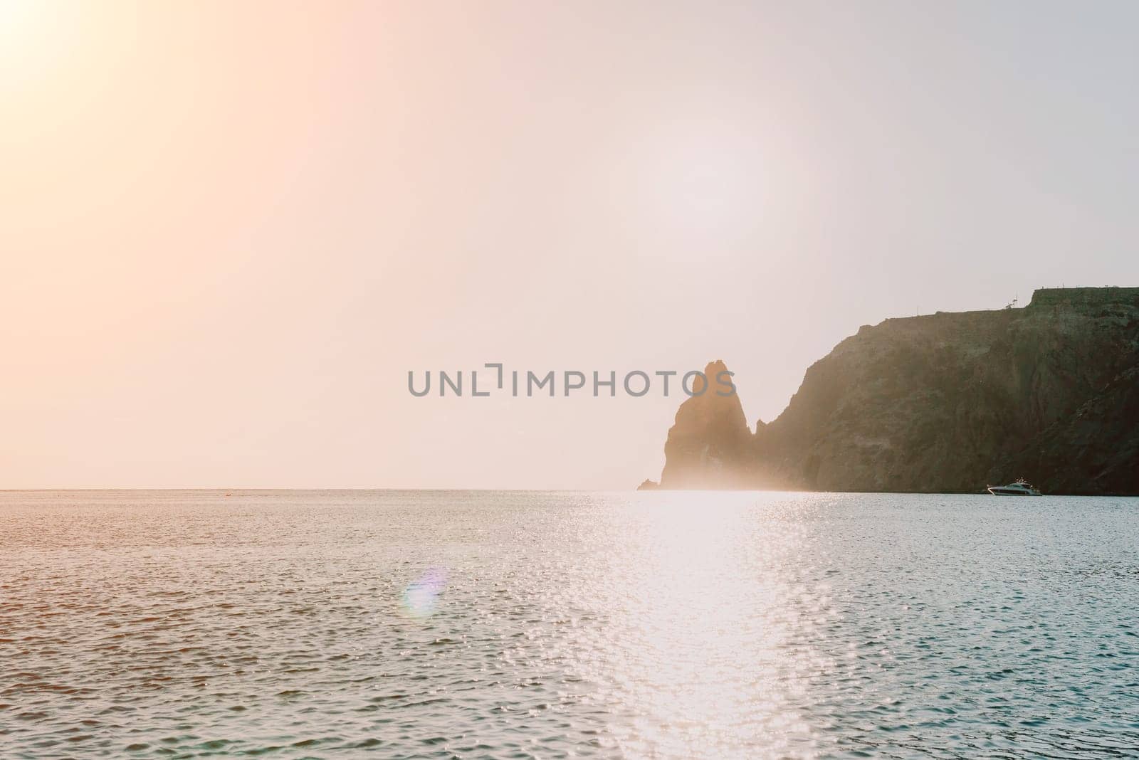 A red burning sunset with the silhouette of a cliff over the sea. Abstract nature summer or spring ocean sea background. Small waves on golden warm water surface with bokeh lights from sun.