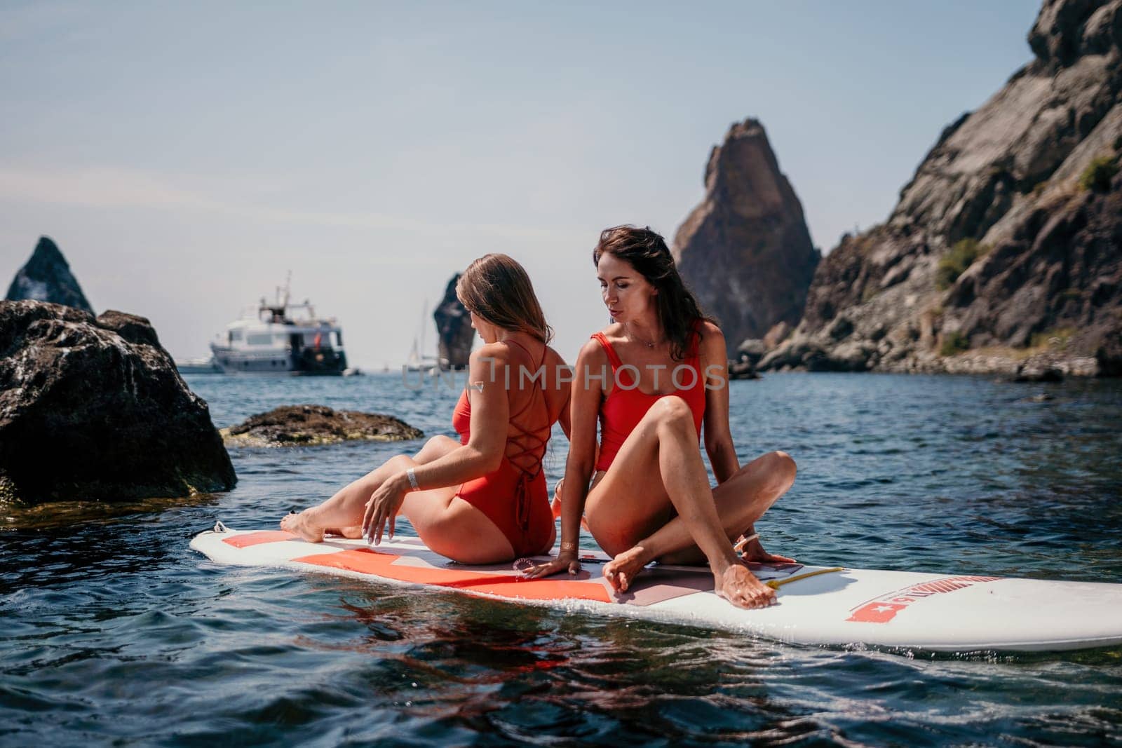 Woman sup yoga. Two happy sporty women practising yoga pilates on paddle sup surfboard. Female stretching doing workout on sea water. Modern individual female hipster outdoor summer sport activity. by panophotograph
