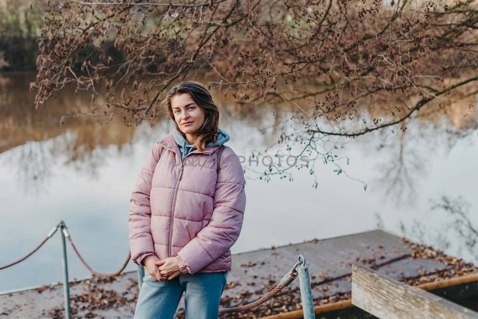 A young woman standing at the shore looking at the river in autumn sunny day. Street view, copy space for text, travel photo. Happy tourist woman on the bank of the river in autumn in warm clothes. Tourists enjoy their vacation, winter season. Romantic look and travel concept. A joyful mood in a caucasian girl. Winter Wonderland: Enchanting Girl by the Riverside in Autumn by Andrii_Ko