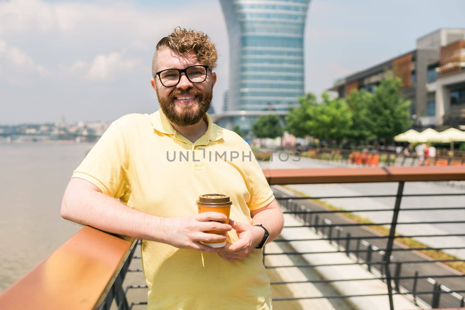Attractive young millennial man with curly hair wearing glasses is walking the street with a cup of coffee on a sunny day. Bearded hipster guy wearing casual clothes enjoys relaxing on a summer weekend. Belgrade city and copy space by Satura86