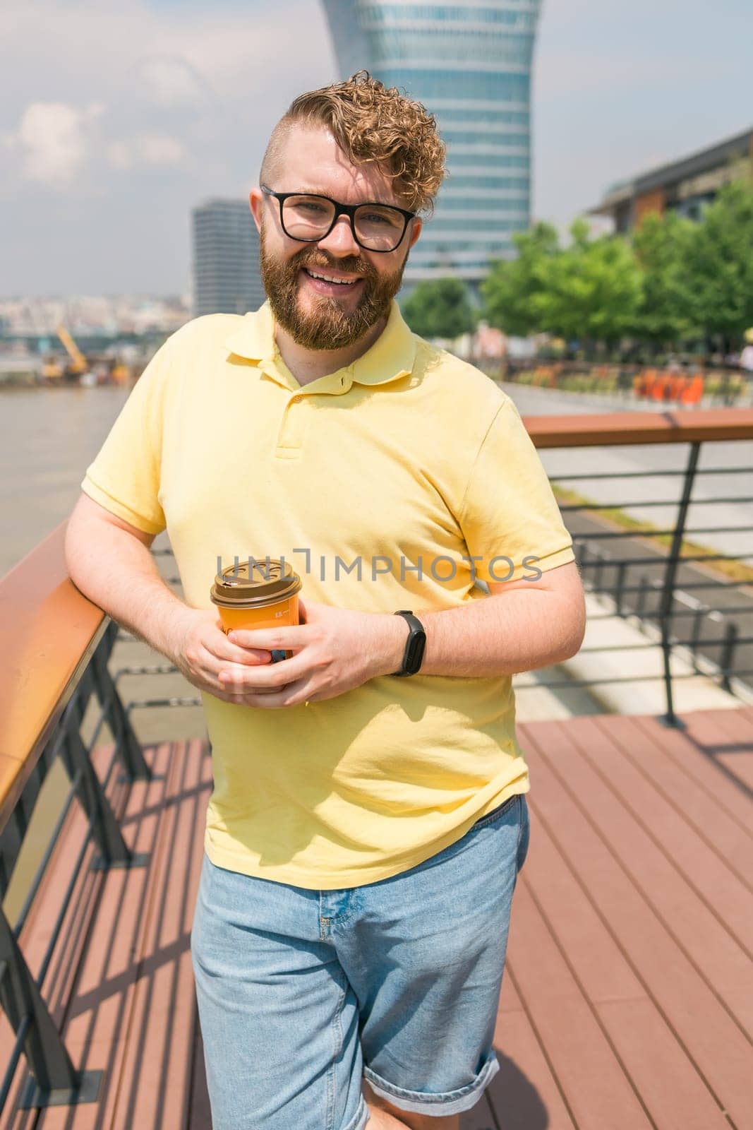 Attractive young millennial man with curly hair wearing glasses is walking the street with a cup of coffee on a sunny day. Bearded hipster guy wearing casual clothes enjoys relaxing on a summer weekend. Belgrade city by Satura86