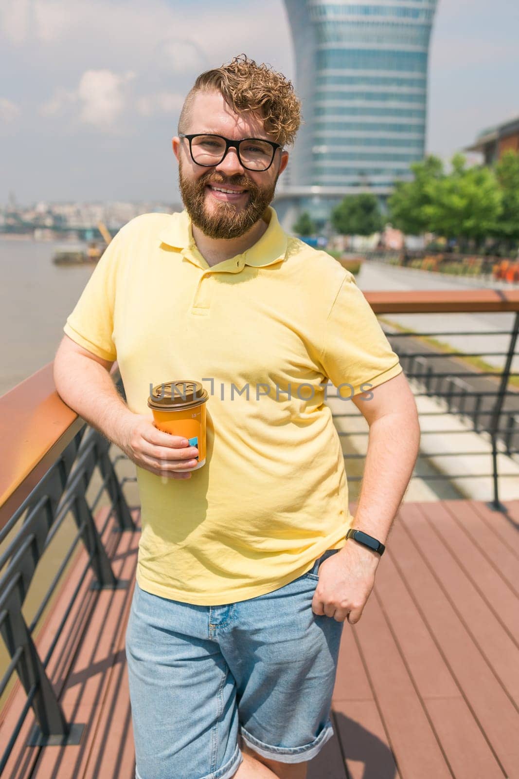 Attractive young millennial man with curly hair wearing glasses is walking the street with a cup of coffee on a sunny day. Bearded hipster guy wearing casual clothes enjoys relaxing on a summer weekend. Belgrade city and copy space.