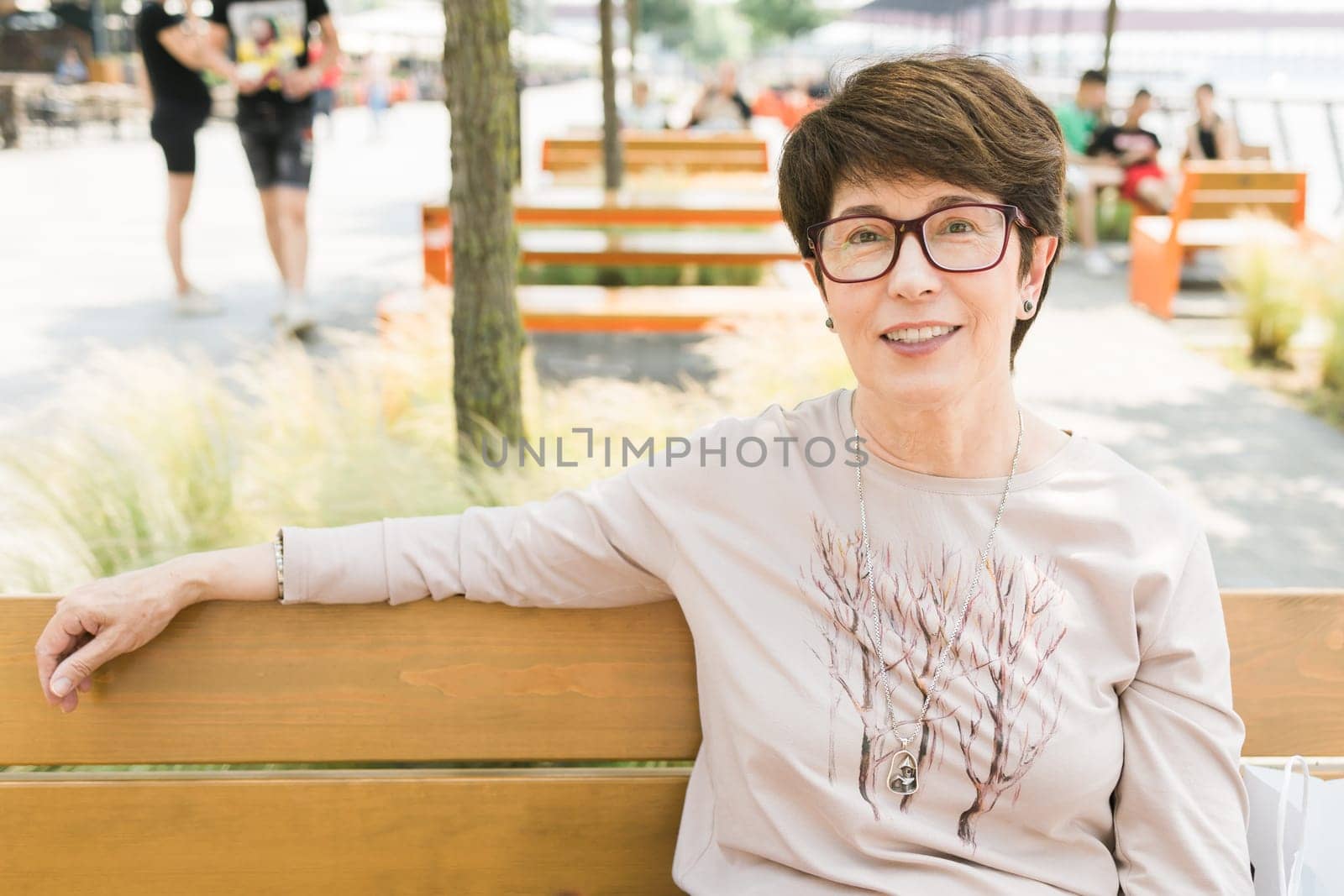 Outdoor portrait of beautiful and elegant middle age 55 - 60 year old woman wearing long sleeve top sitting on bench on the street. Copy space and empty place for text advertising.