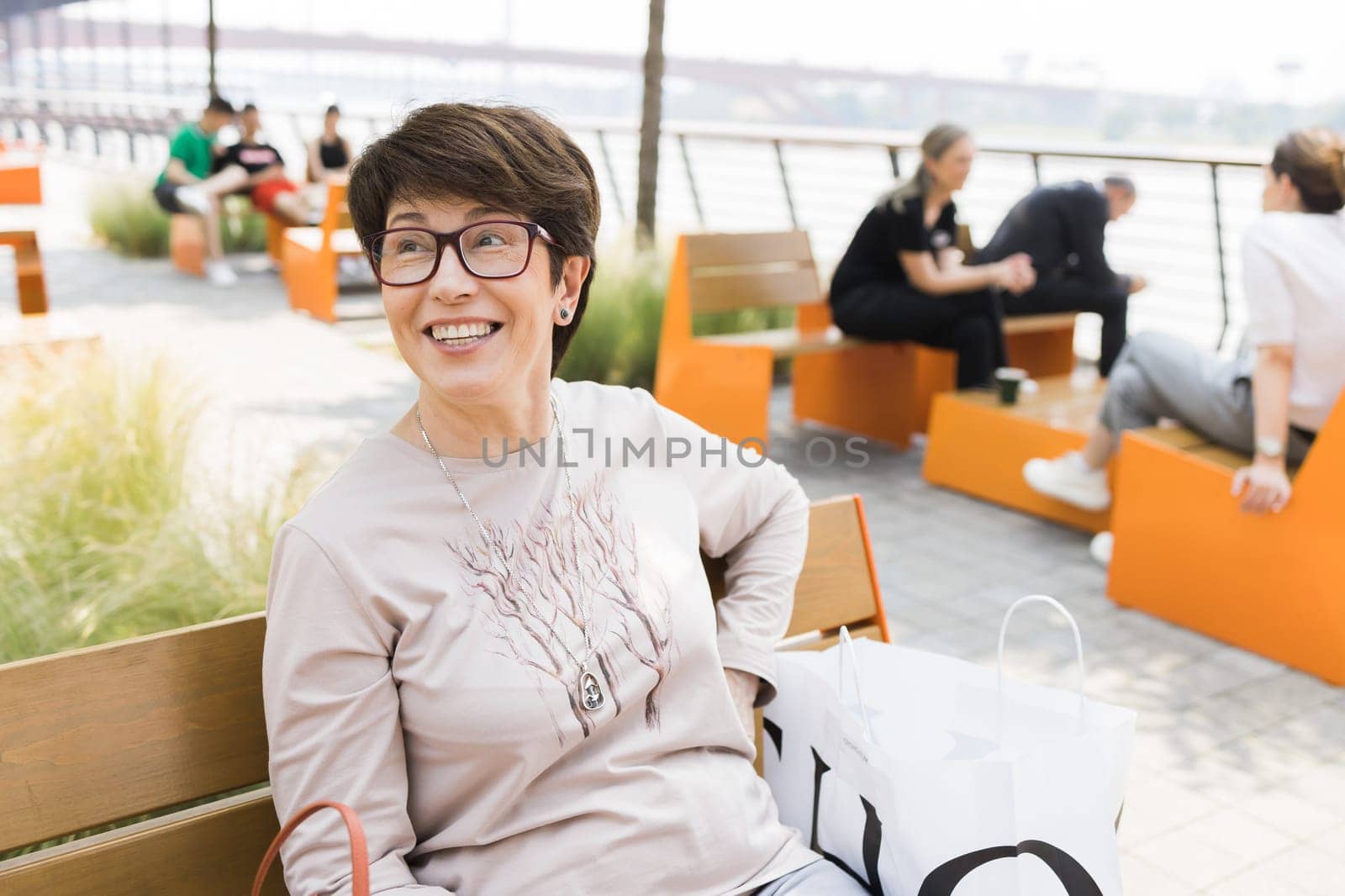 Outdoor portrait of beautiful and elegant middle age 55 - 60 year old woman wearing long sleeve top sitting on bench on the street. Copy space and empty place for text advertising.