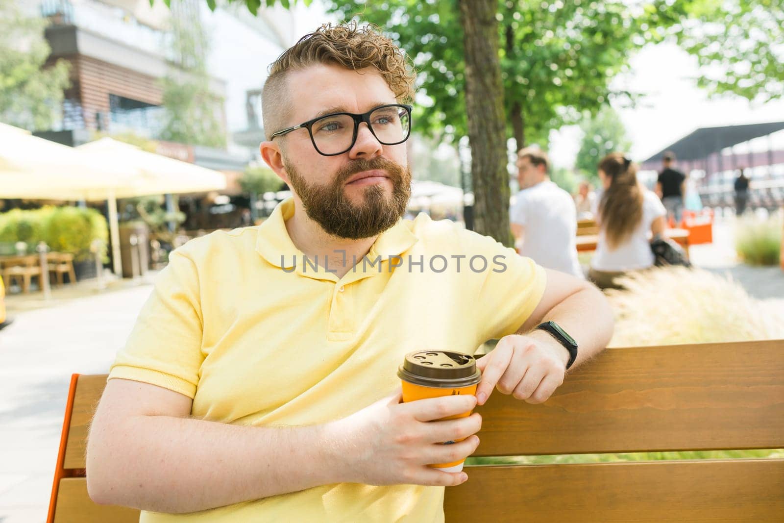 Millennial smiling student man in yellow polo t-shirt eyeglasses sitting on bench drink coffee have breakfast rest relax in sunshine spring green city park outdoors on nature. Urban leisure concept