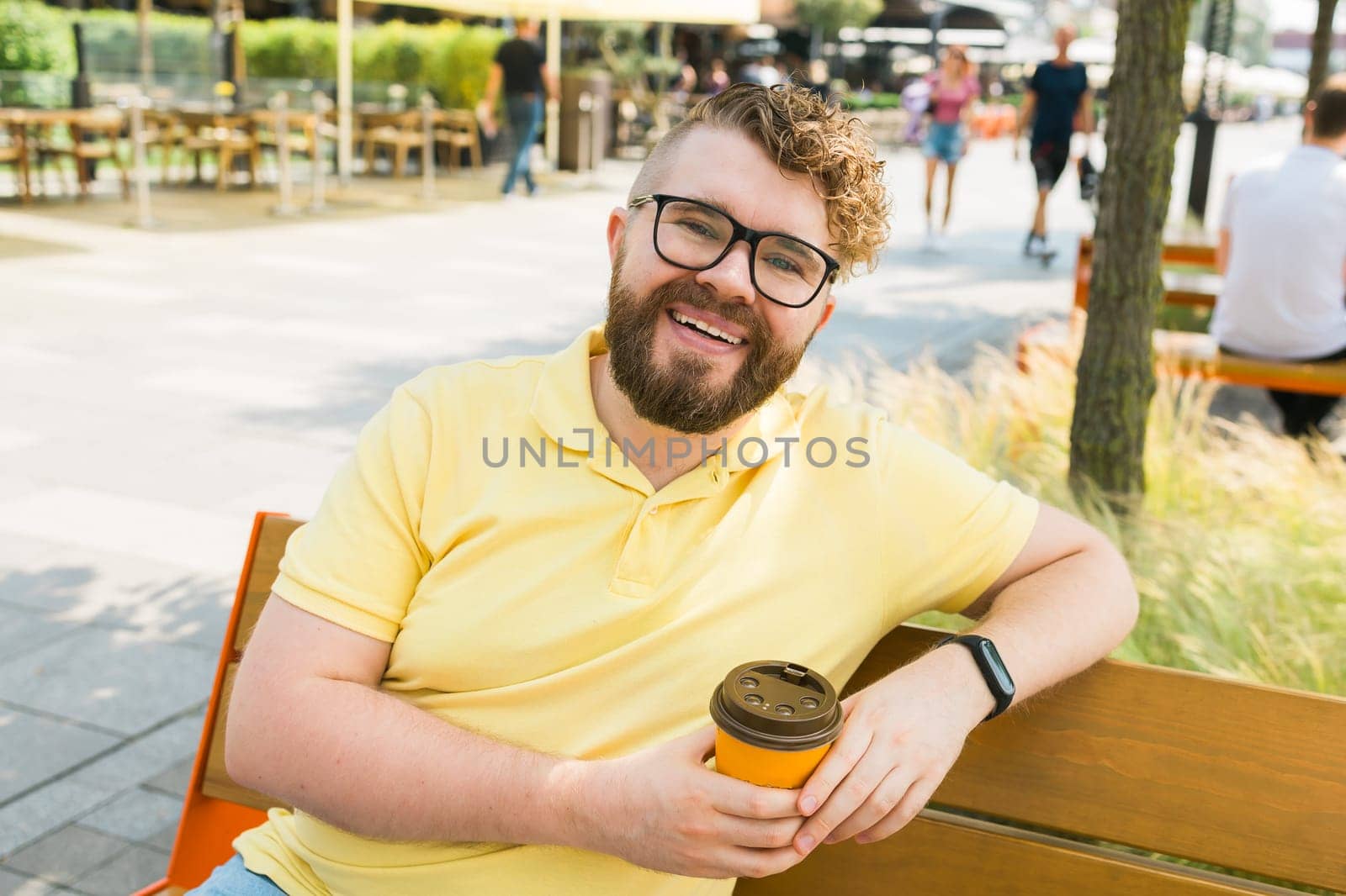 Millennial smiling student man in yellow polo t-shirt eyeglasses sitting on bench drink coffee have breakfast rest relax in sunshine spring green city park outdoors on nature. Urban leisure concept
