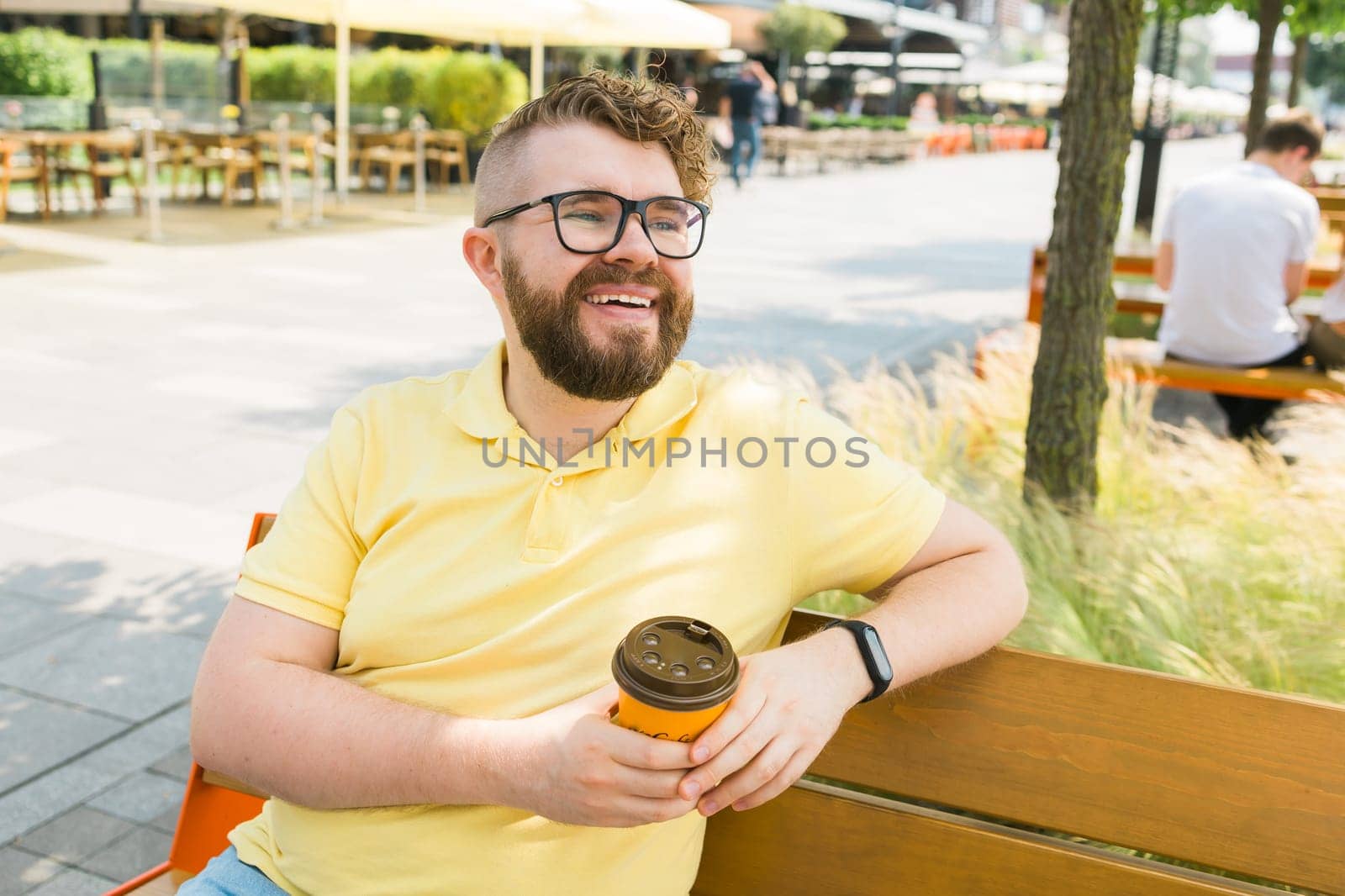 Millennial smiling student man in yellow polo t-shirt eyeglasses sitting on bench drink coffee have breakfast rest relax in sunshine spring green city park outdoors on nature. Urban leisure concept. Copy space by Satura86