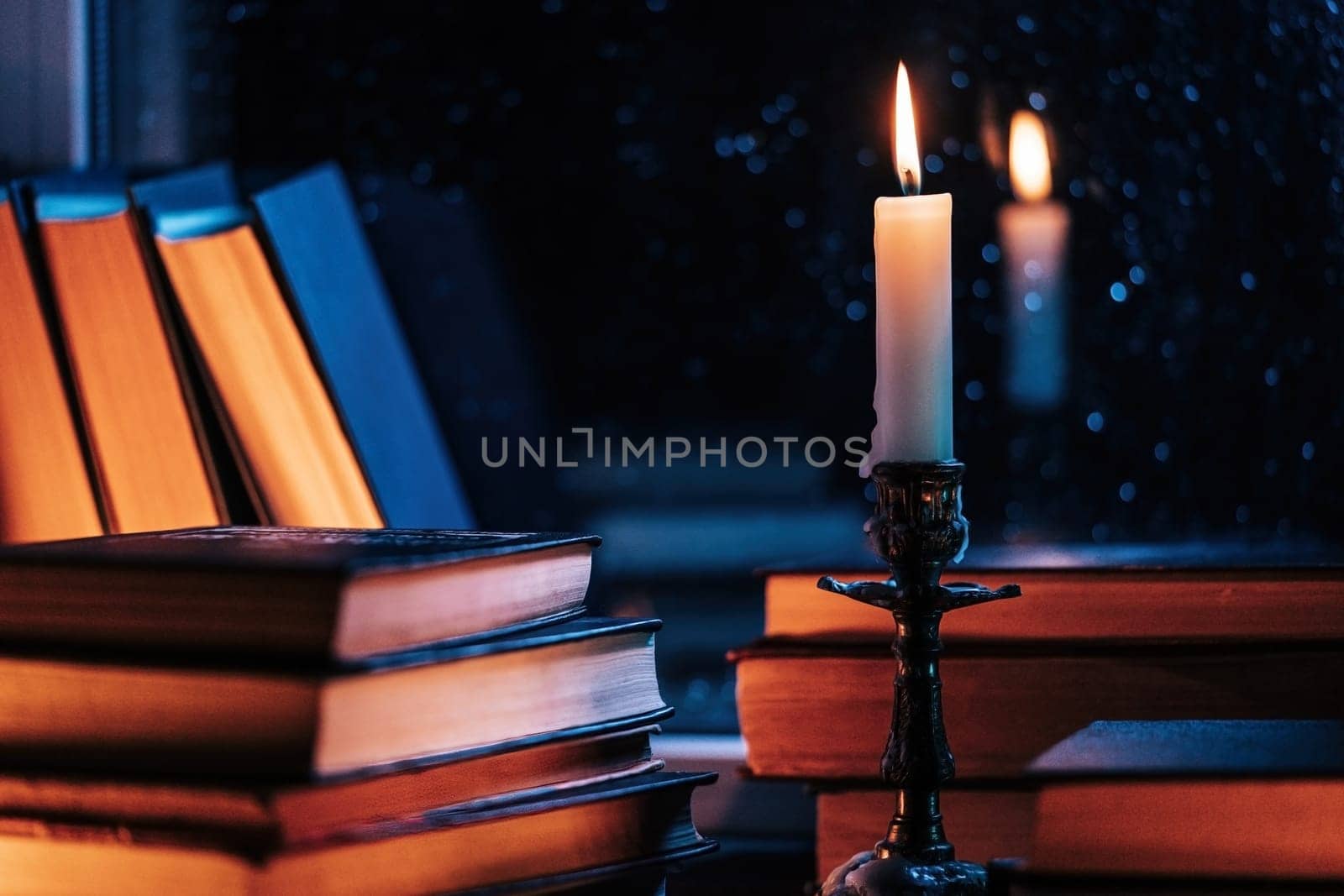 Stack of old paper books on wooden windowsill, bathed in soft candle glow light. Cozy ambience of fall, candle burning. Literature promotions or tranquil visual storytelling. Rainy autumn weather.