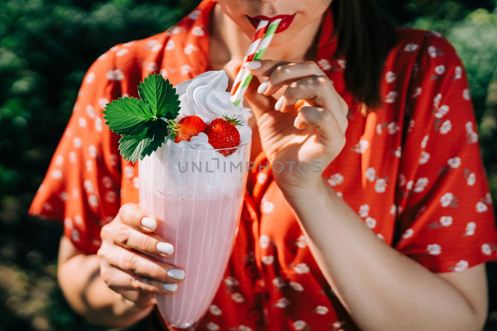 Pretty woman drinking strawberry milkshake or smoothie cocktail with straw, nature backdrop. Appetizing summer dessert. Healthy berry food. High quality