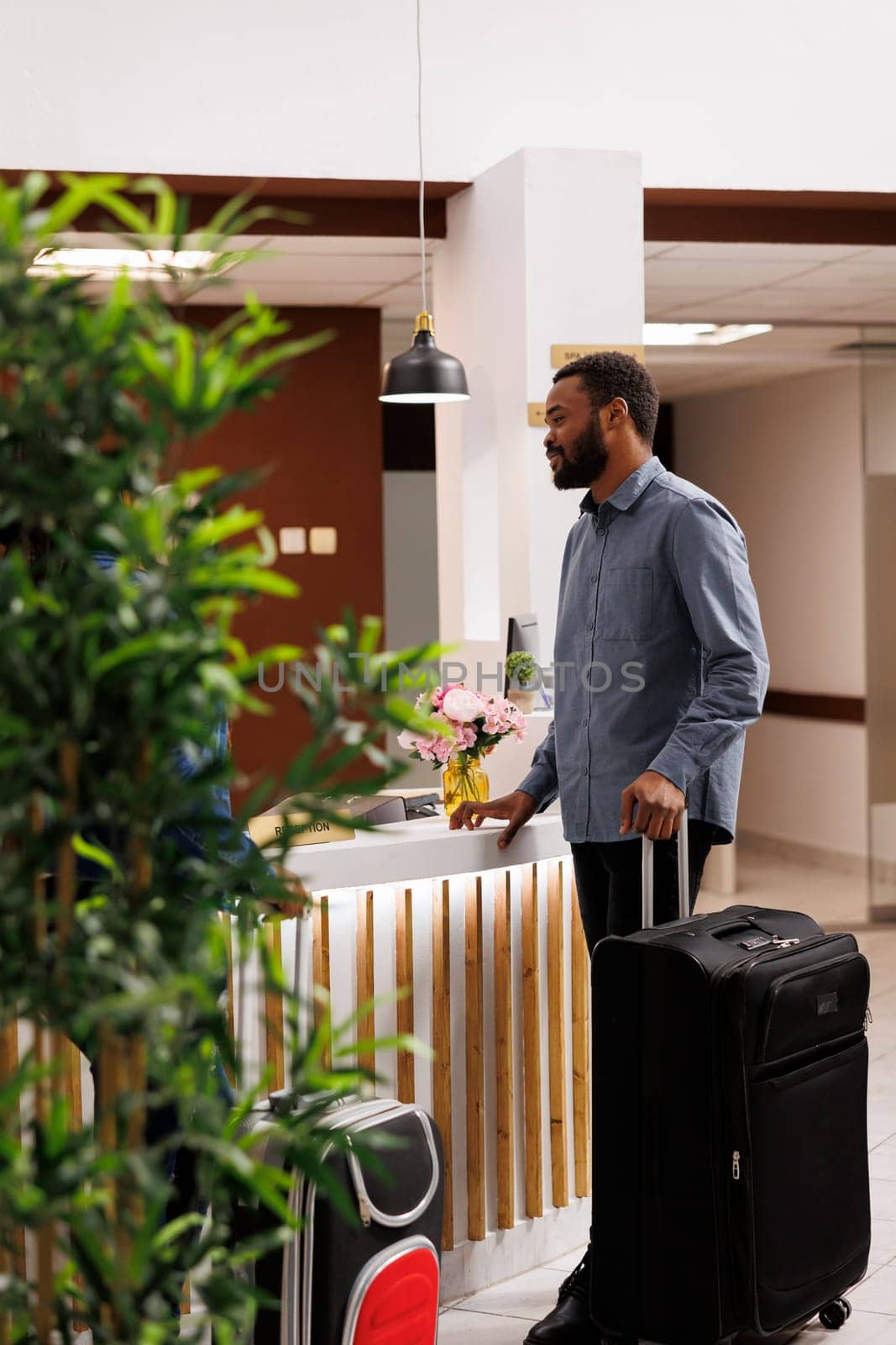 Young African American man with suitcase standing at reception area, talking pleasantly with front desk employees, waiting in line to check-in after arrival. Black guy tourist checking out of hotel