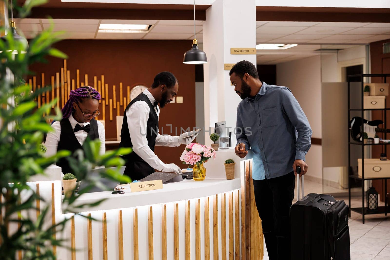 Front desk staff checking guest passport and travel documents during hotel check-in procedure. African American man receptionist using computer software, tourist with suitcase at reception