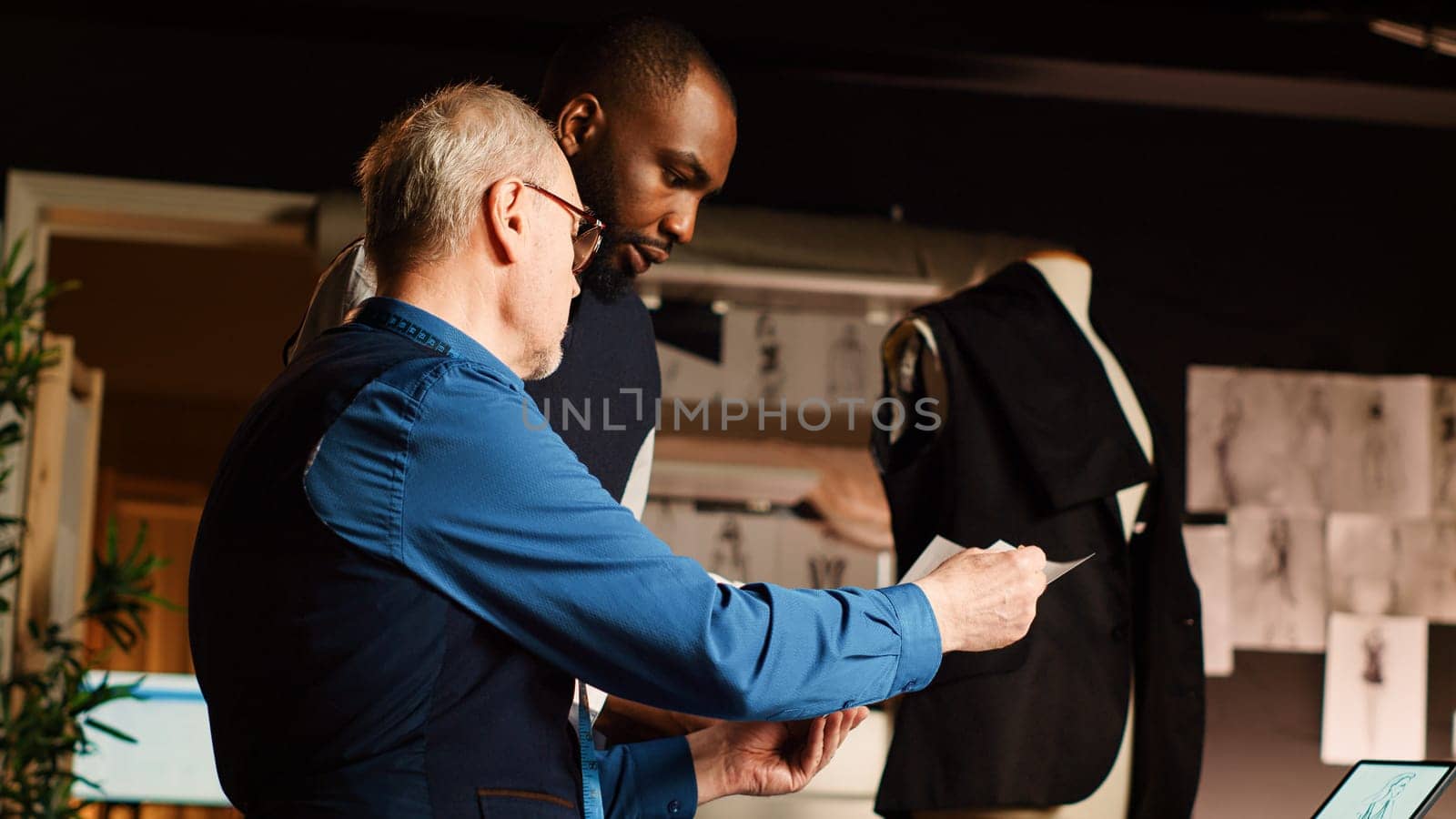 Couturier and pupil checking sketches in tailor workshop, learning to craft clothing line handmade. Fashion designer and apprentice discussing about models and fabric. Handheld shot.