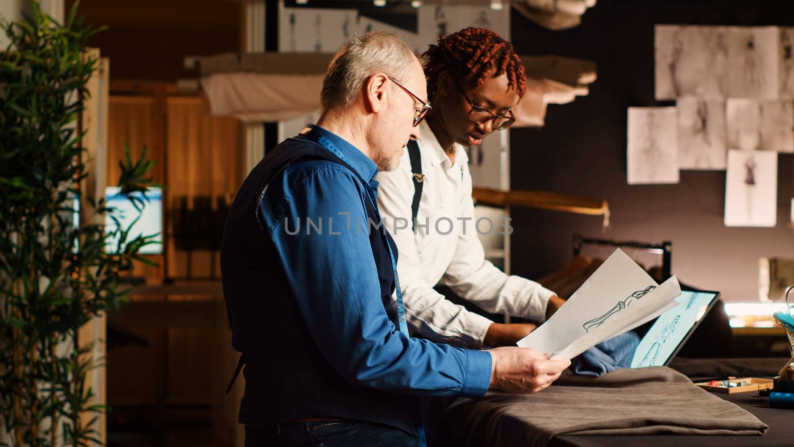 Experienced tailor and student working in atelier, looking at clothes design sketches to craft fashion collection. Apprentice learning the art of sewing from senior couturier. Handheld shot.