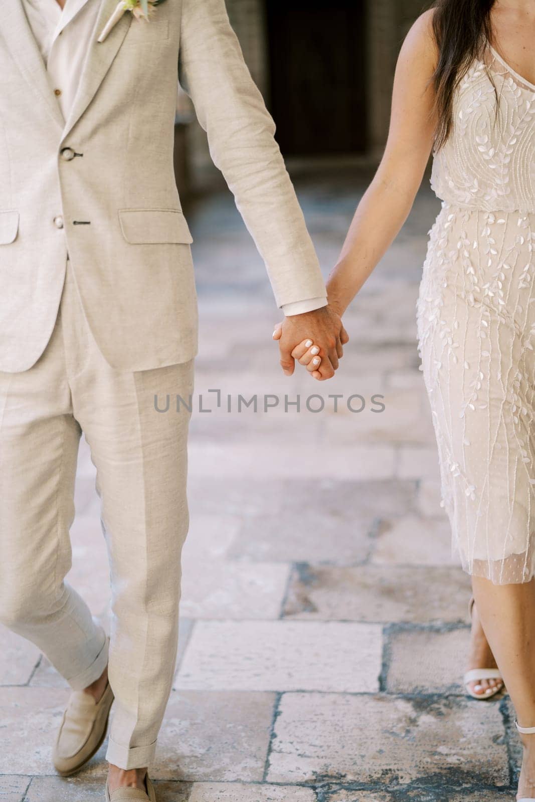 Bride and groom walk holding hands along the paving stones. Cropped. Faceless by Nadtochiy