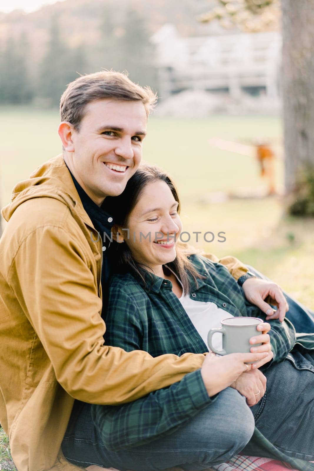 Smiling man hugging woman with mug sitting in autumn park. High quality photo
