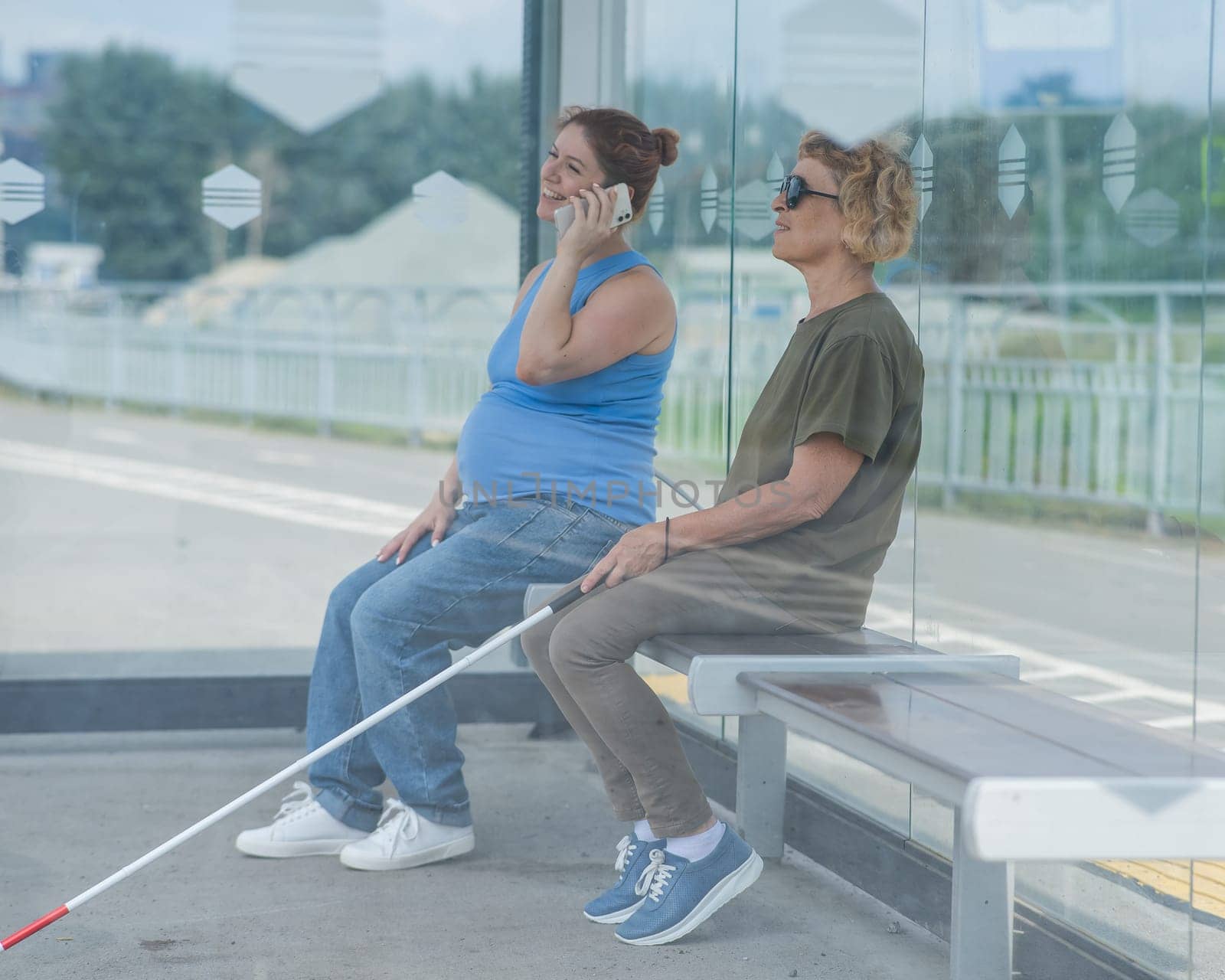 An elderly blind woman and a pregnant woman are sitting at a bus stop and waiting for the bus. by mrwed54