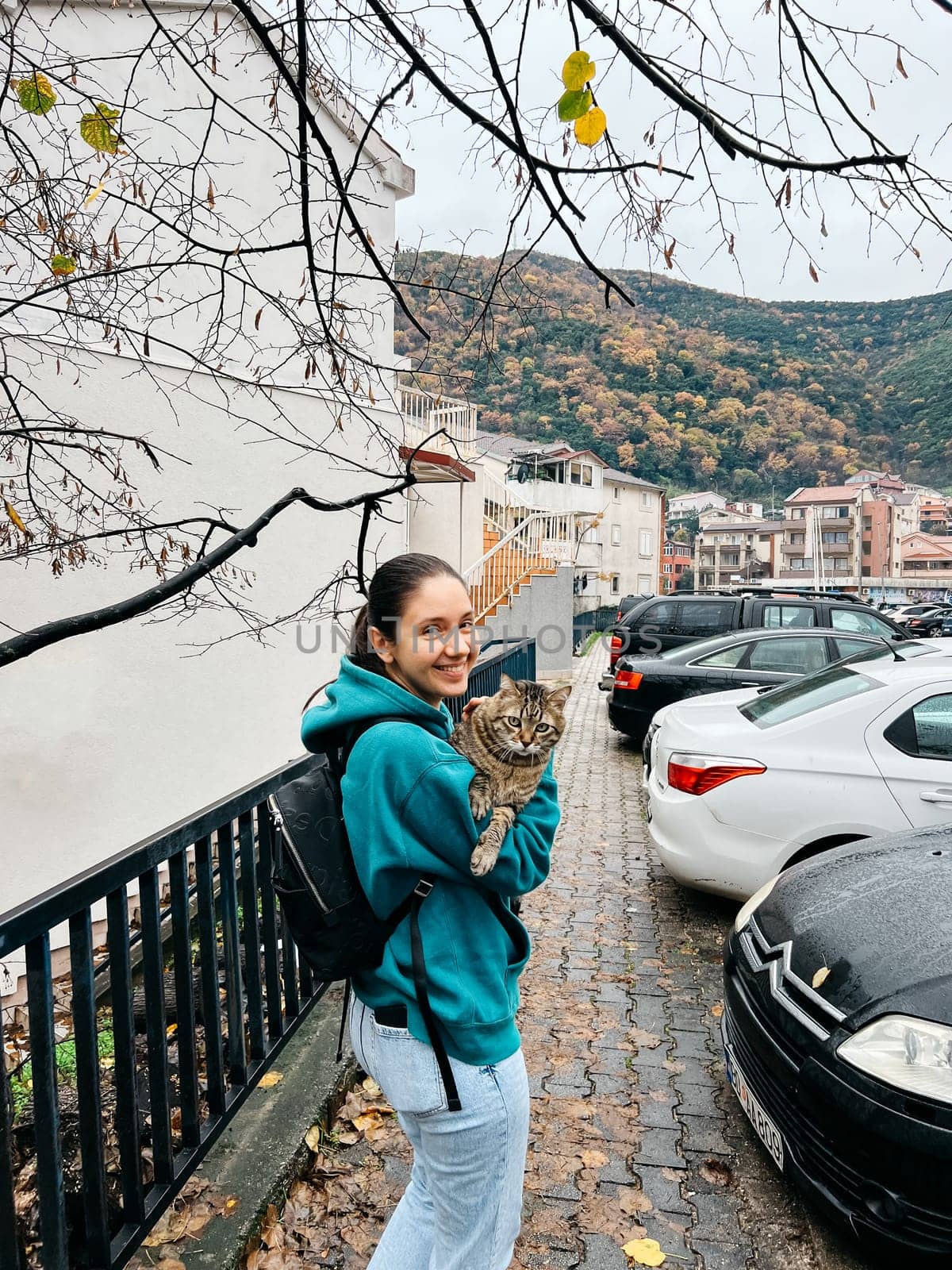 Smiling young woman with a big cat in her arms stands near the house on the street. High quality photo