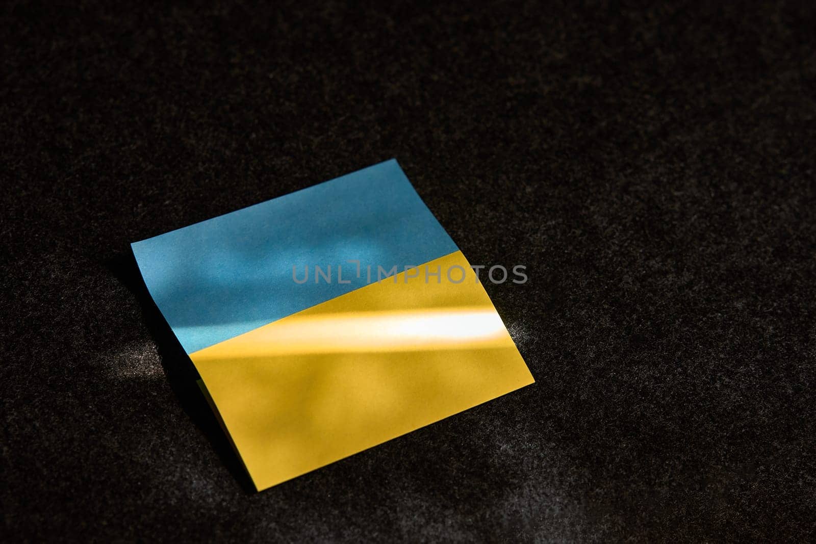 The sticker in the form of the Ukrainian flag lies. on a dark surface and a ray of sunlight falls on it by snep_photo