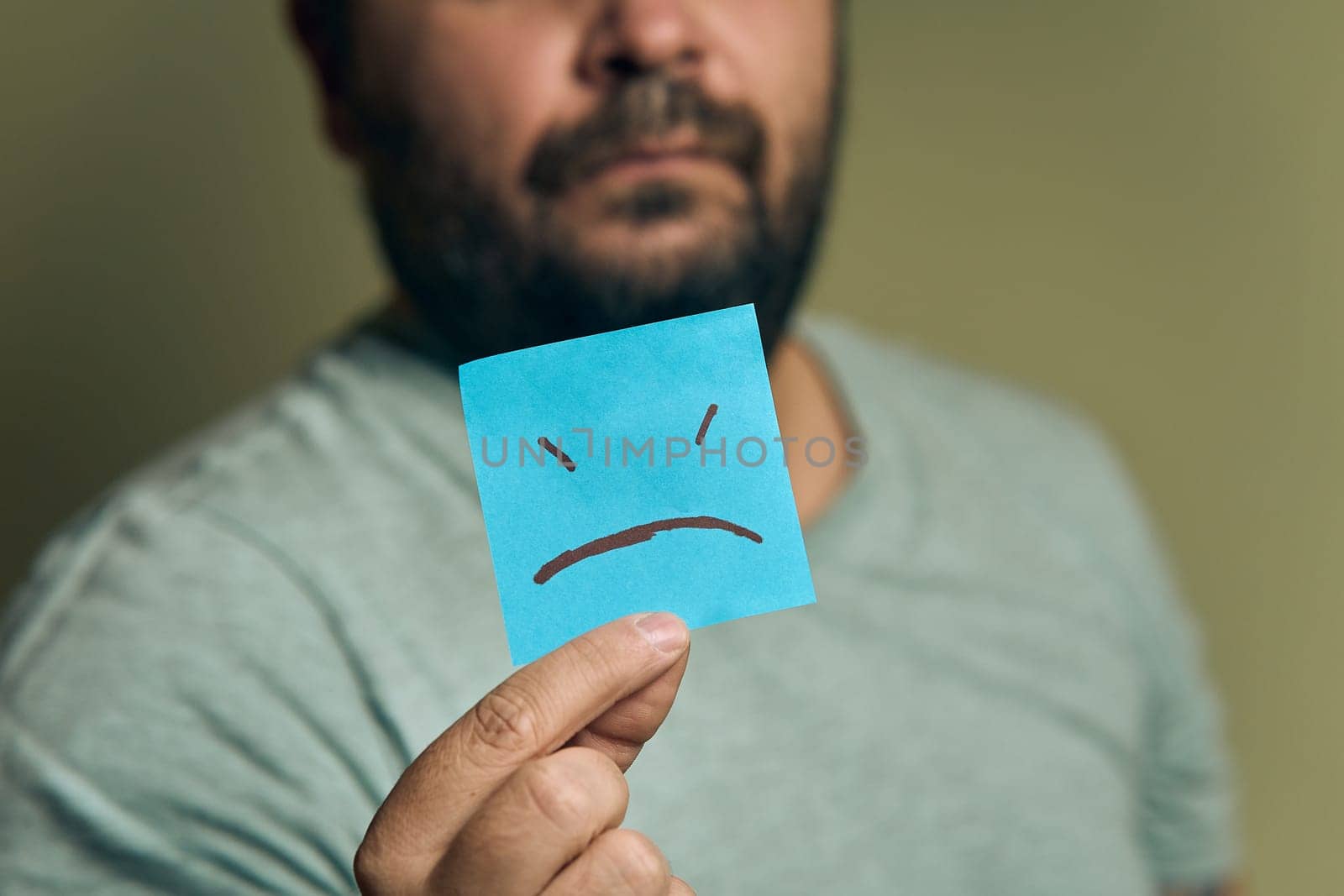 A bearded European man holds a orange sticker in front of him, with a sad smiley