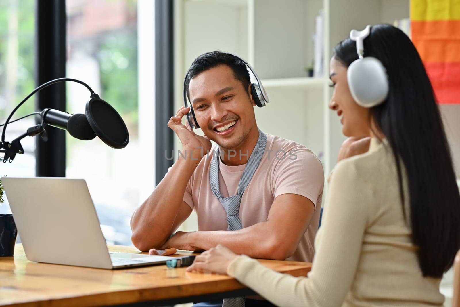 Positive gay male radio host discussing various topics with guest while streaming live audio podcast in studio.