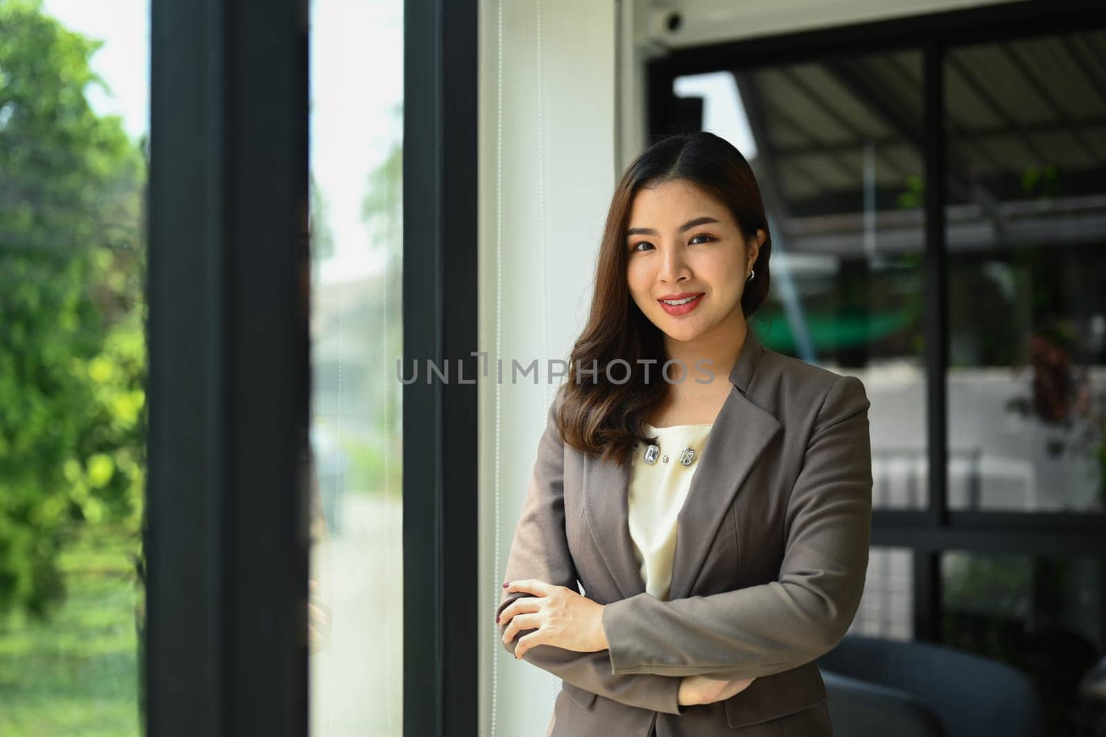 Portrait of successful millennial businesswoman in luxury suit standing with arms crossed and smiling at camera.